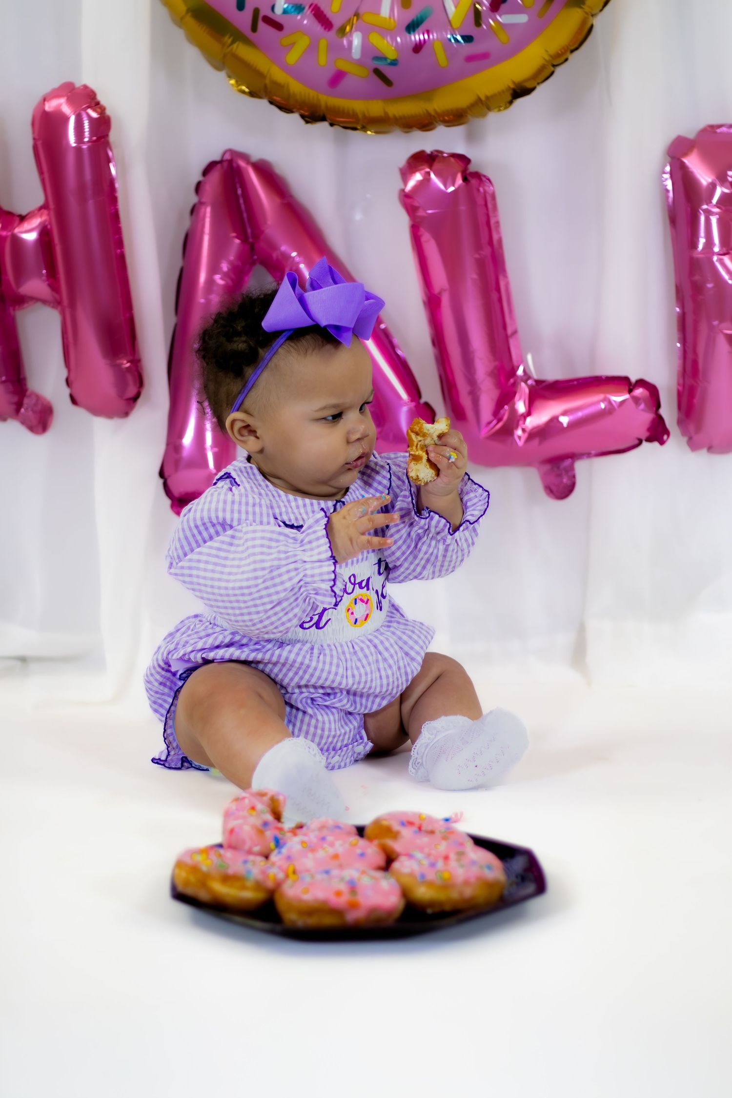 Adorable baby in purple outfit eats donut at half birthday party with pink balloons.