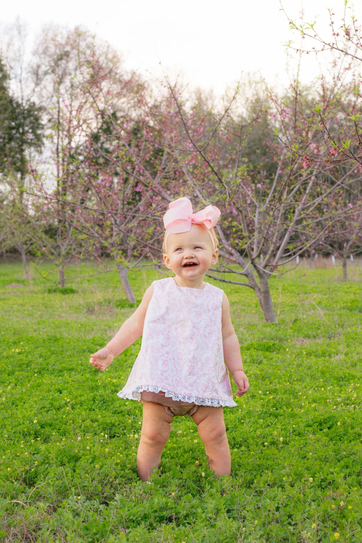Laughing baby girl in white lace dress and pink bow stands on green grass among blooming spring trees.