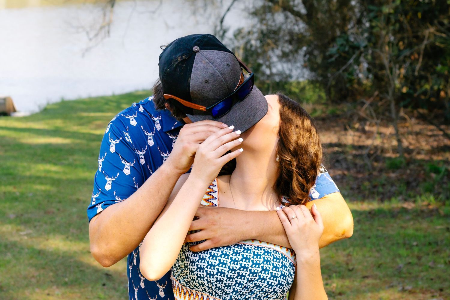 Couple sharing a romantic kiss outdoors near lake, man in blue deer print shirt embracing woman.