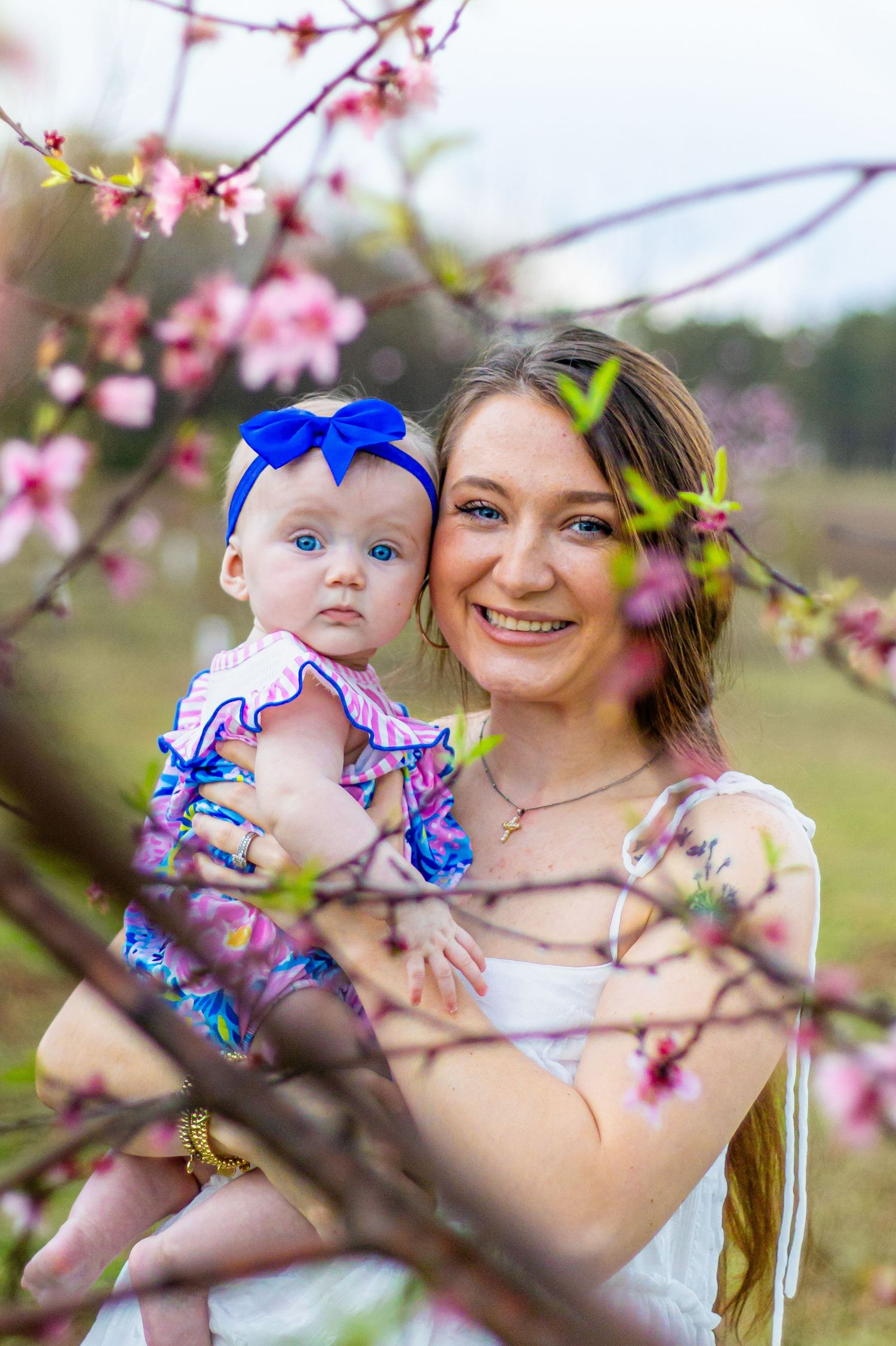 Smiling mother holds baby girl with blue bow amid pink cherry blossoms in spring orchard.