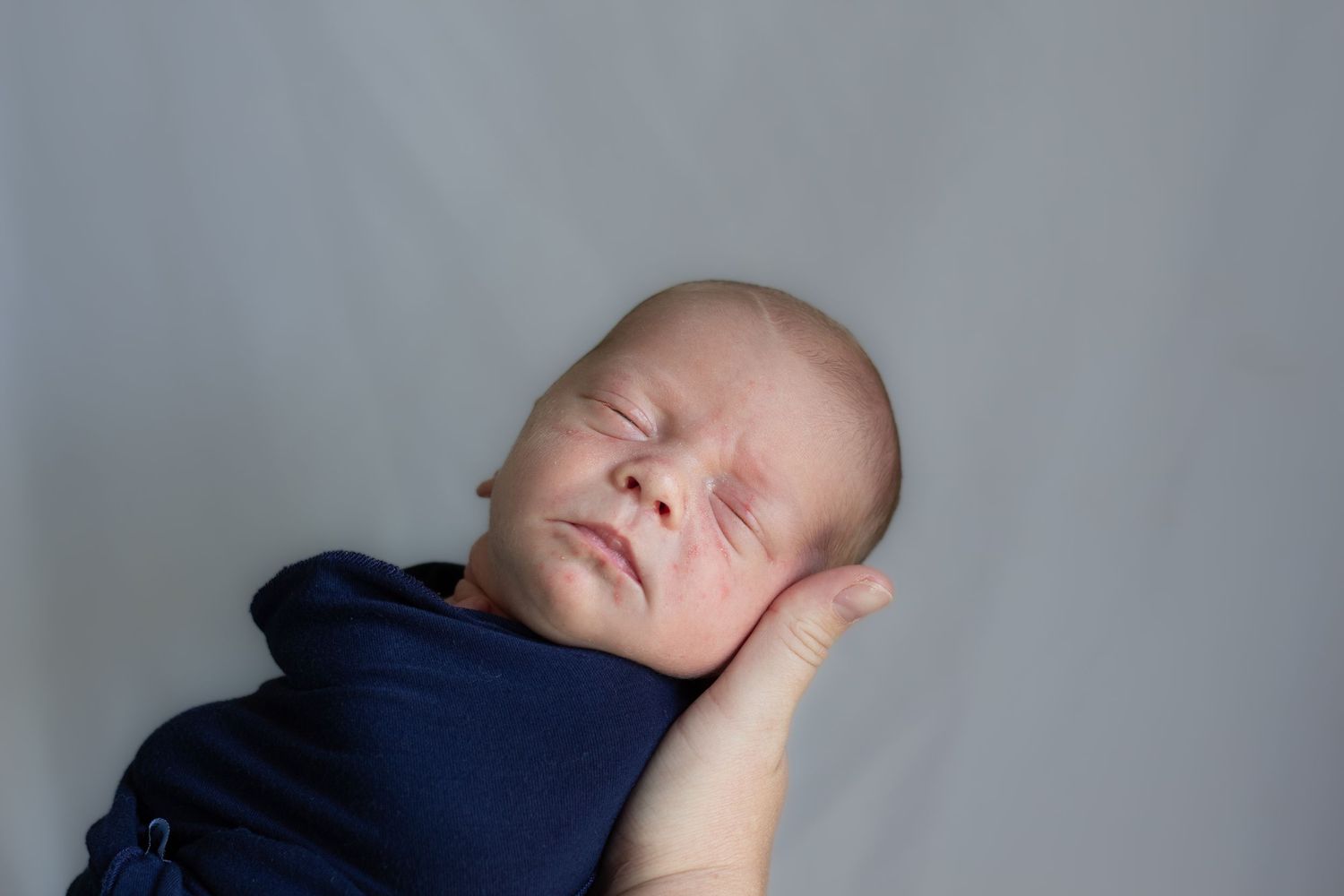 Peaceful sleeping newborn baby swaddled in navy blue wrap on gray background.