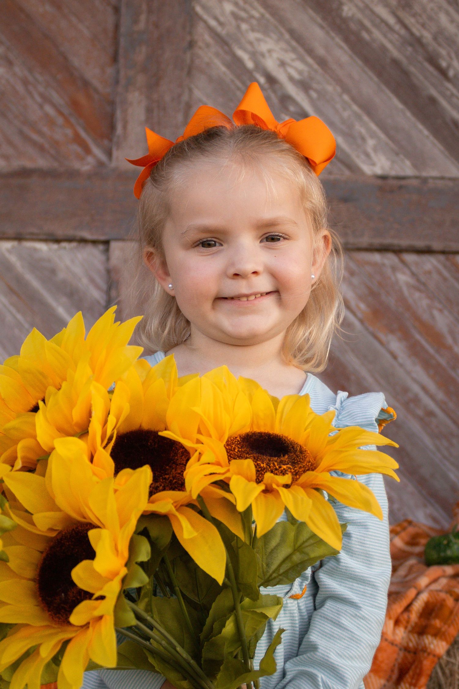 Smiling blonde girl with orange bow holds a large bouquet of yellow sunflowers at a fall photo shoot.