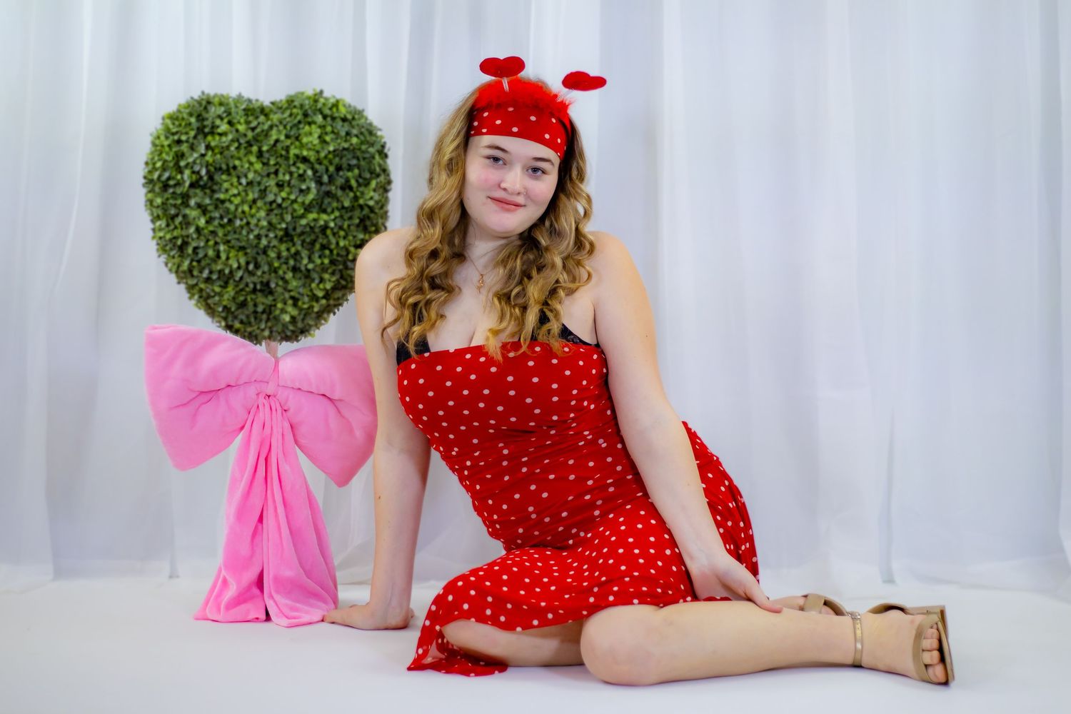 Young woman in red polka dot dress and red headband poses beside heart-shaped topiary with pink bow.