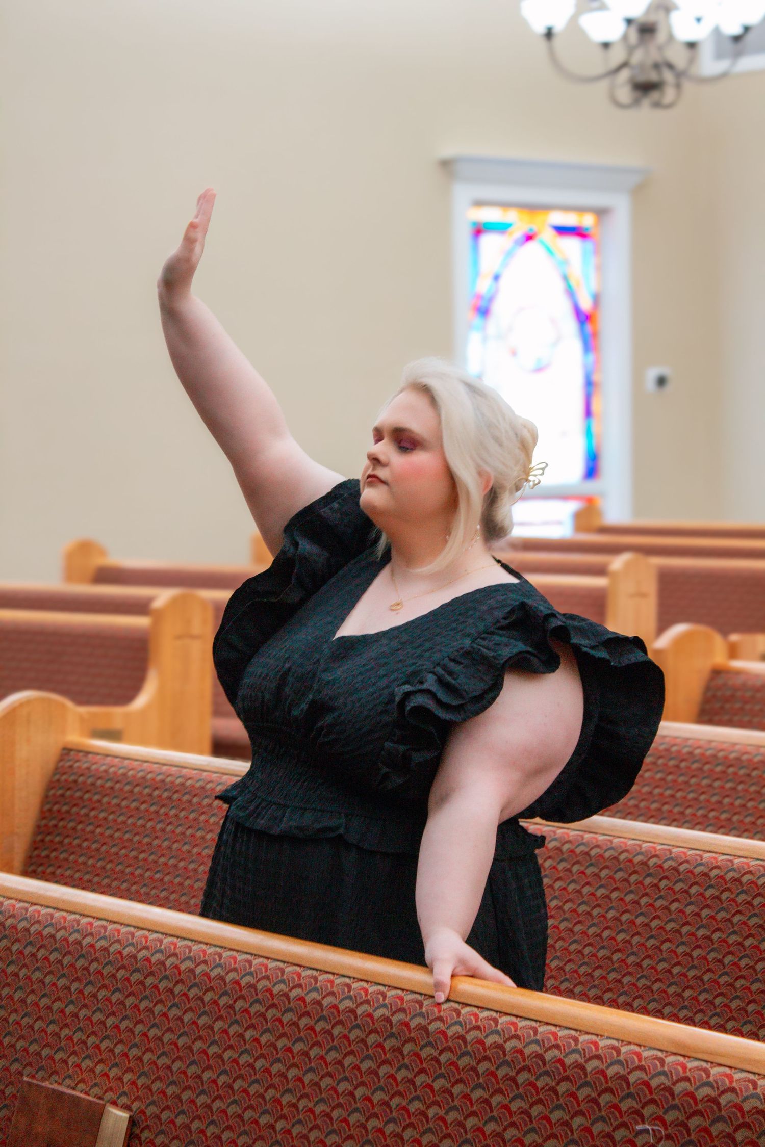A plus-size blonde woman in a black dress raises her hand in worship among church pews with stained glass.