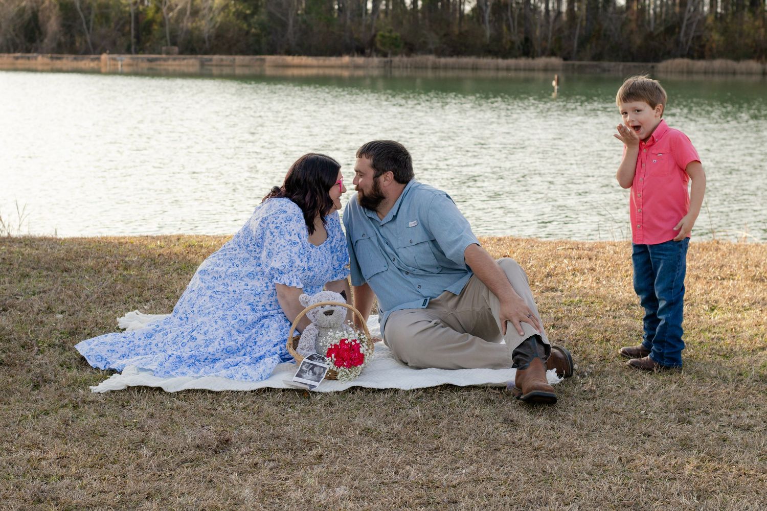 A couple shares a romantic kiss on a blanket by a lake while their young son watches during a maternity shoot.