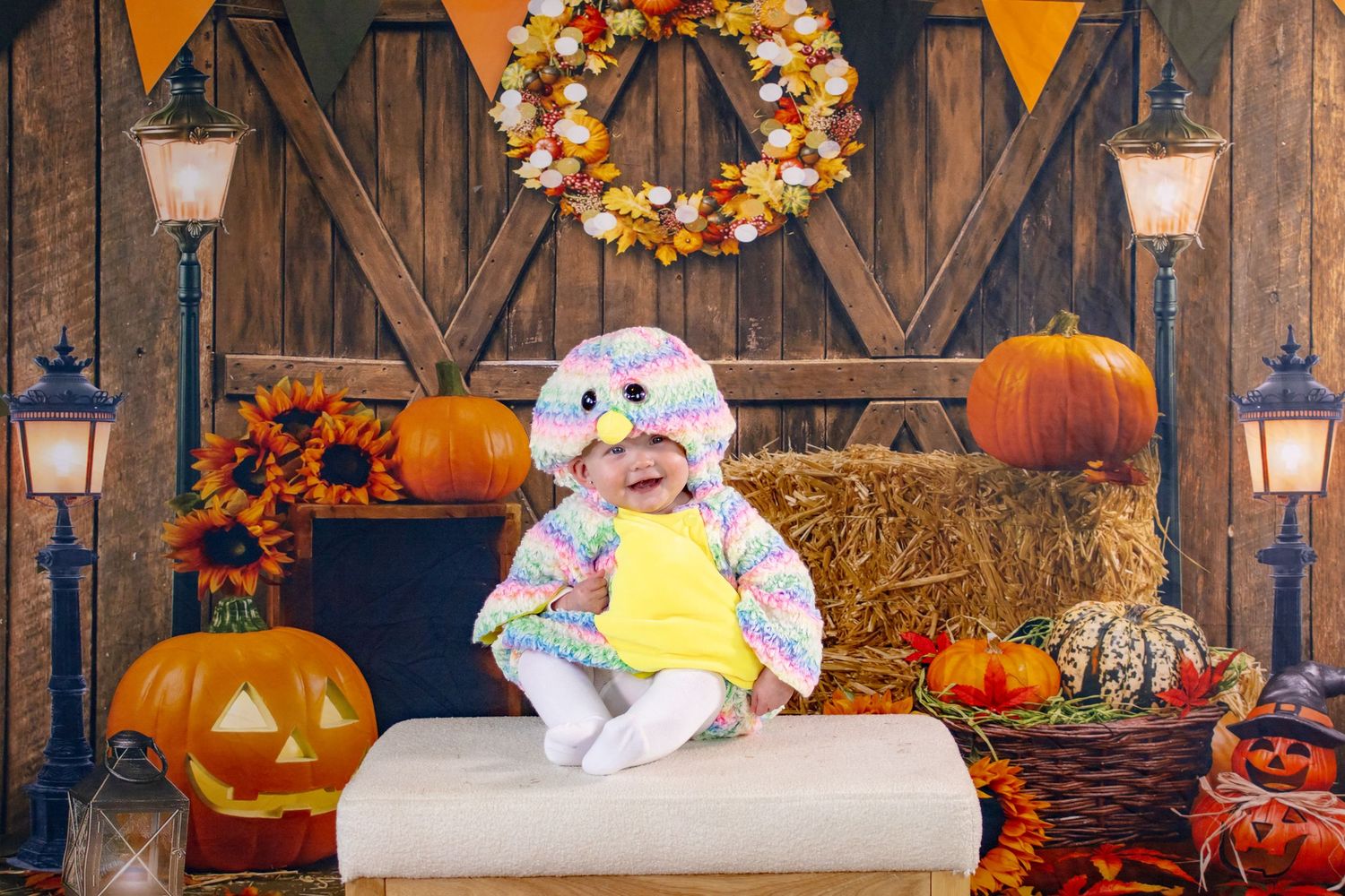 Cute baby in colorful owl costume sits among Halloween pumpkins and fall decorations.