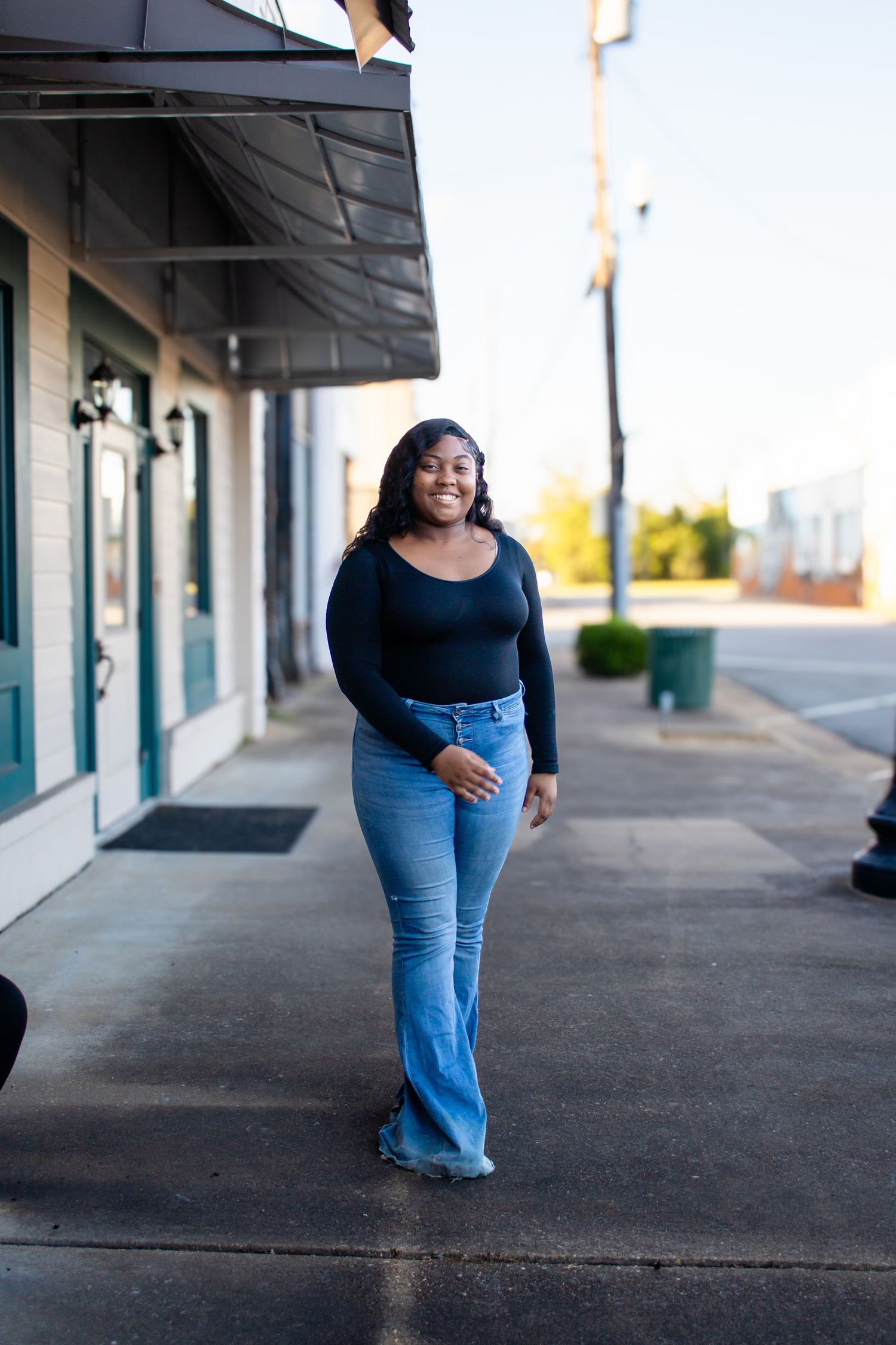 Smiling young Black woman in black long-sleeve top and blue flare jeans walking on urban sidewalk.