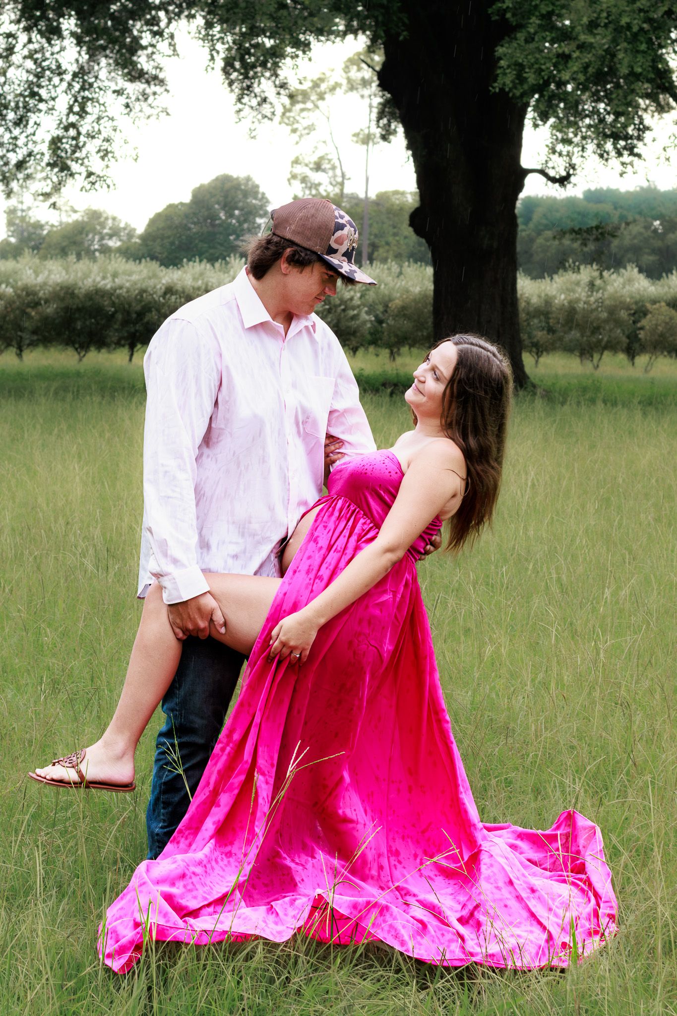 A couple poses romantically in a green field, woman in pink gown lifted by man in white shirt and decorative hat.