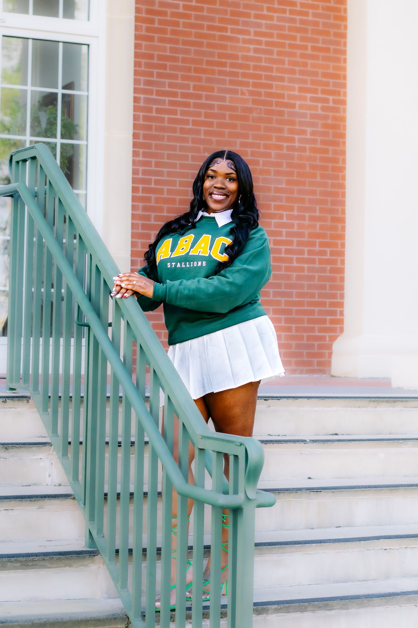 Young woman in ABAC Stallions green sweatshirt and white skirt posing on brick building steps.
