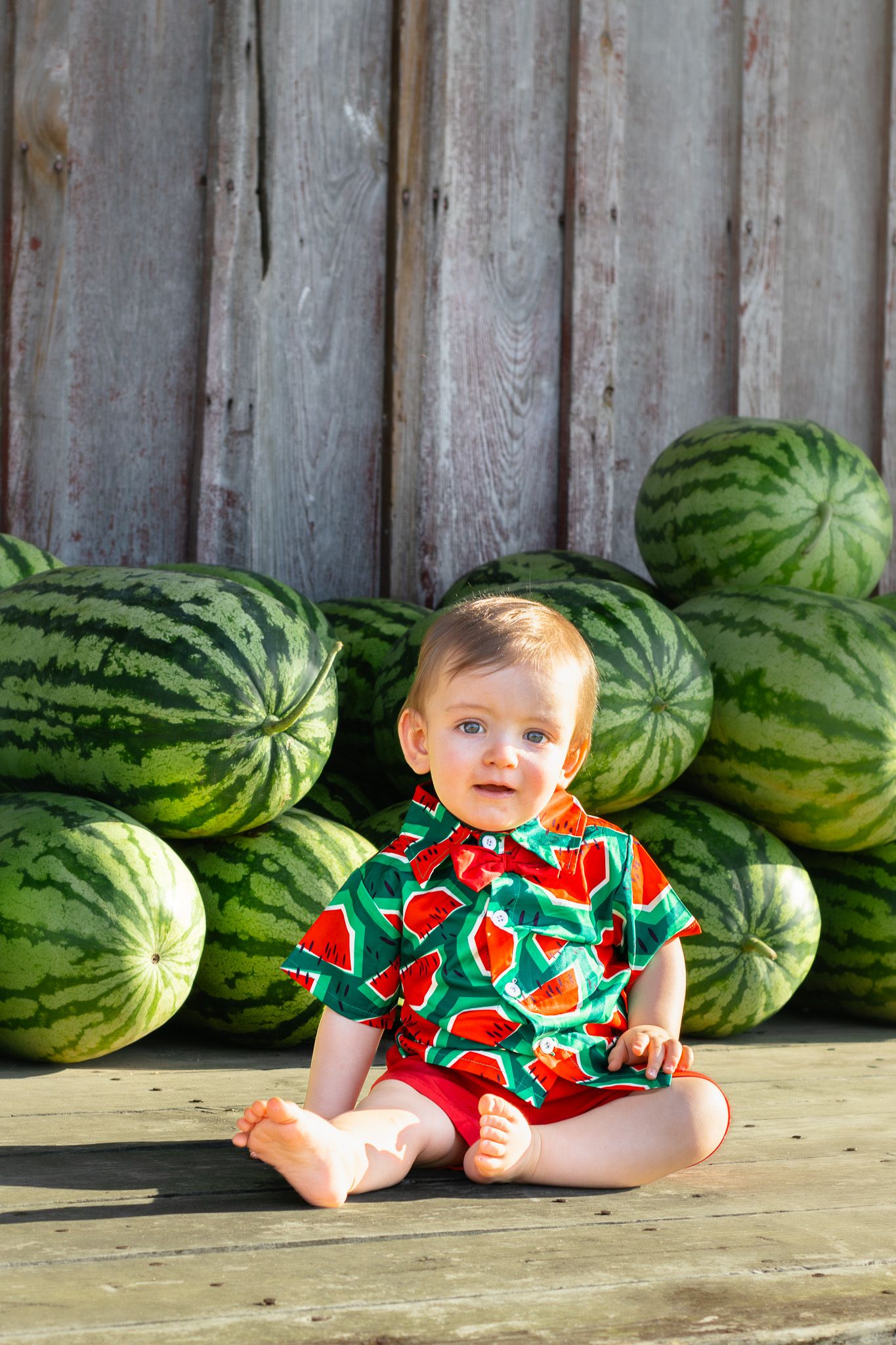 Baby in watermelon print outfit sits cross-legged on wooden deck surrounded by large watermelons.