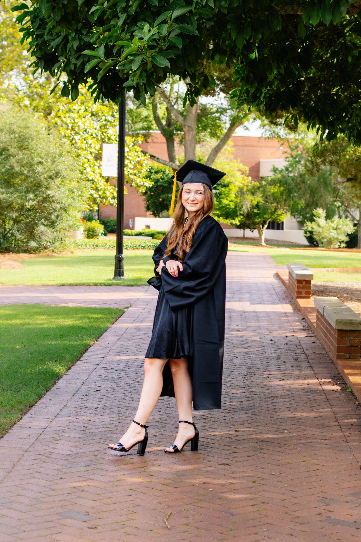 Smiling female graduate poses in black cap and gown on brick pathway surrounded by green trees.