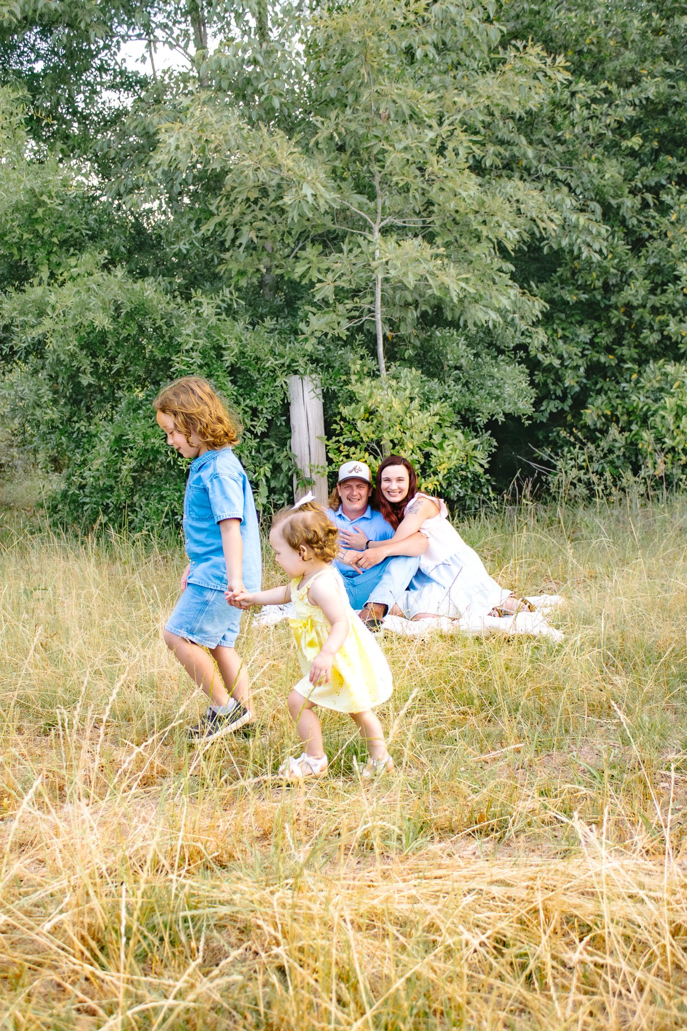 Family enjoying a sunny outdoor picnic in a grassy field with children playing near lush green trees.