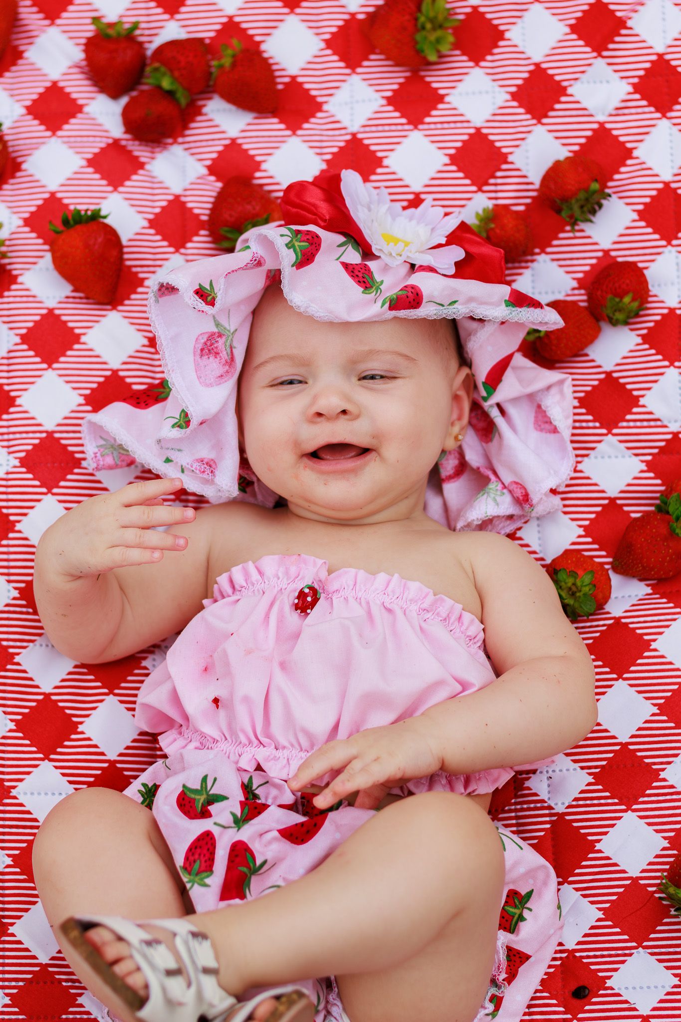 Smiling baby girl in pink strawberry outfit and hat lying on red gingham blanket surrounded by fresh strawberries.