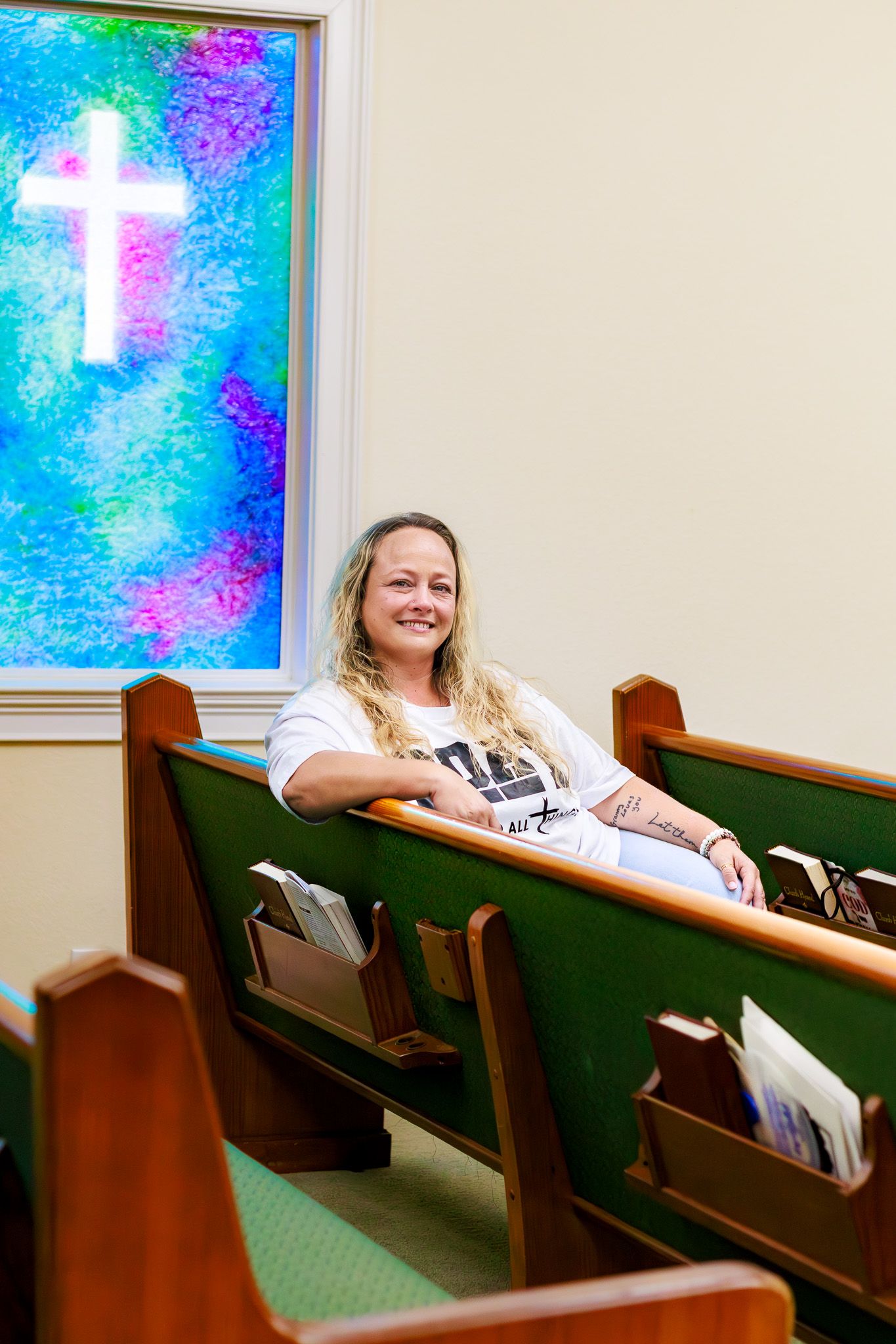 Smiling woman sitting in green church pew near colorful stained glass cross window.