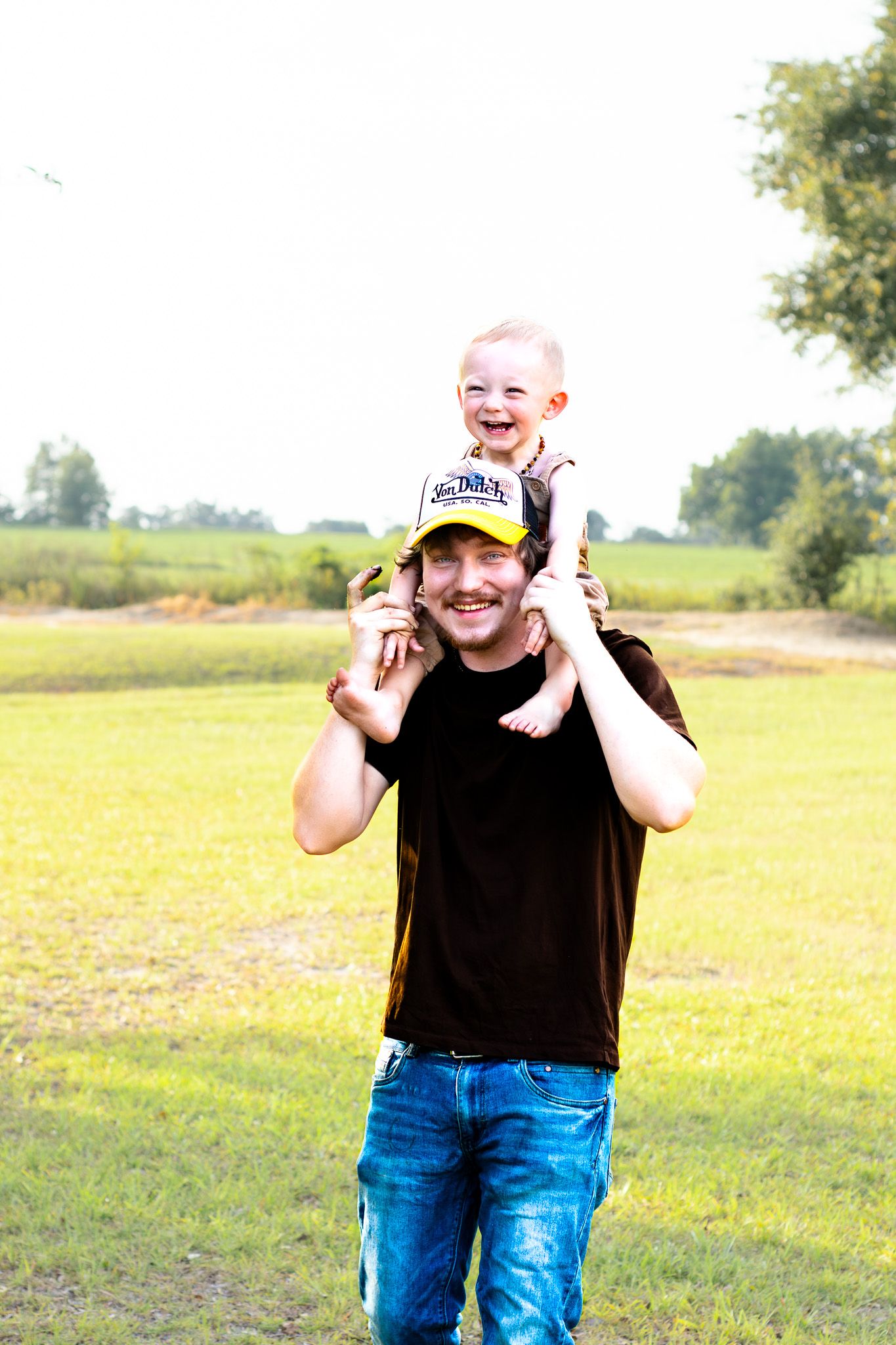 A smiling man in a black shirt carries a laughing toddler on his shoulders outdoors on a sunny day.