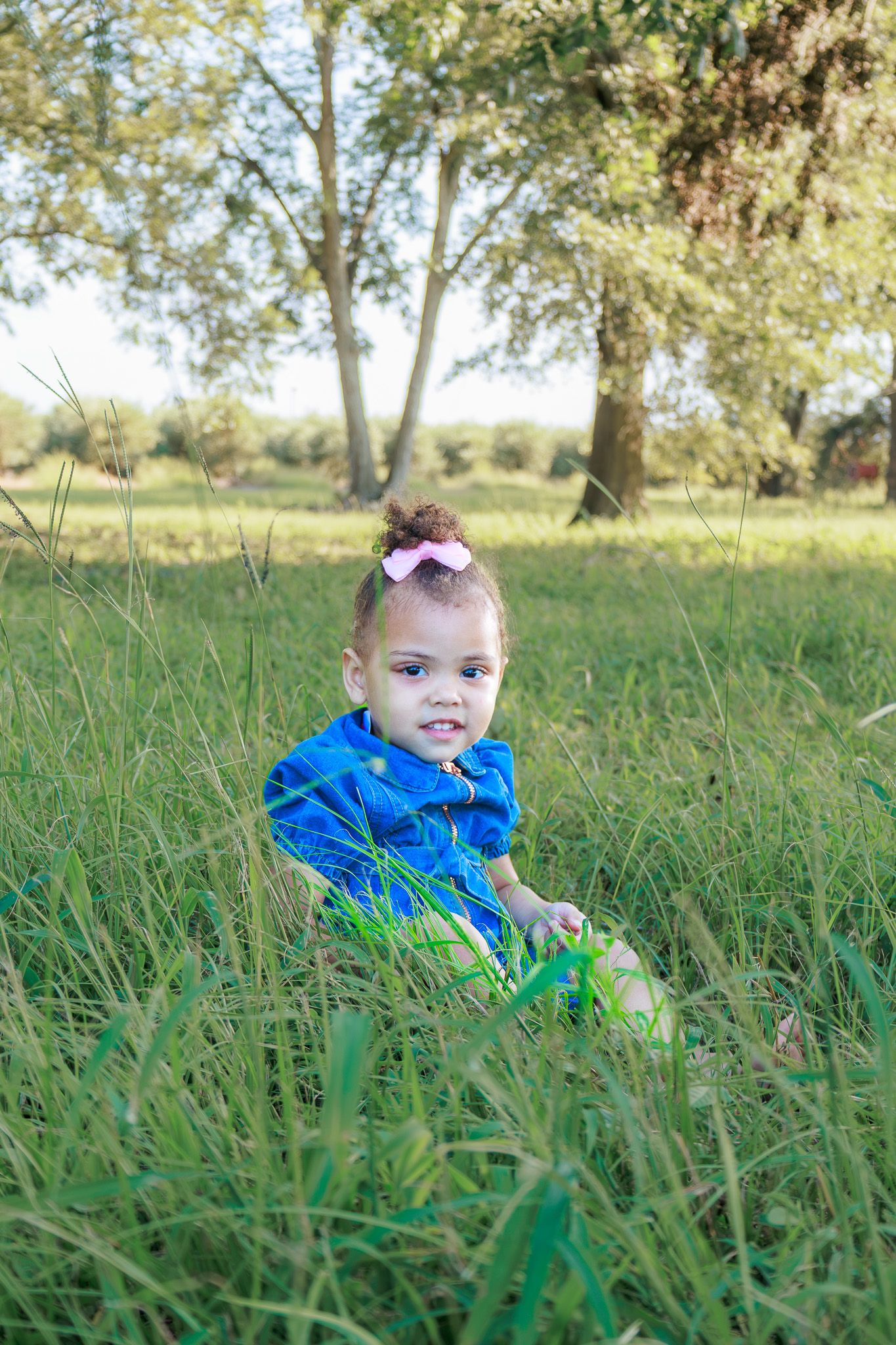 Toddler girl in blue dress with pink bow sits in tall green grass outdoors with trees in background.