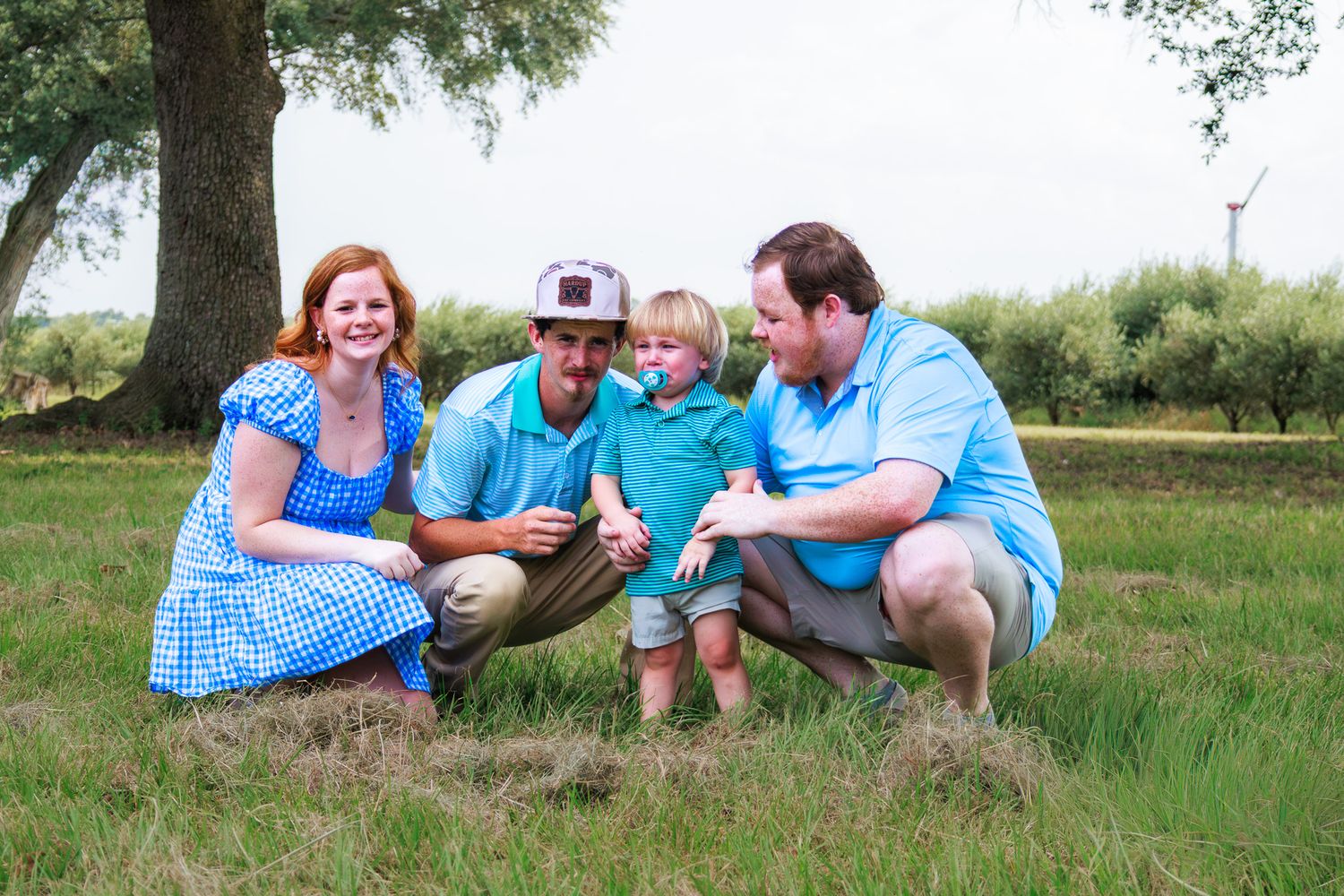 Happy family of four posing in a green field near a tree on a sunny day, smiling together.