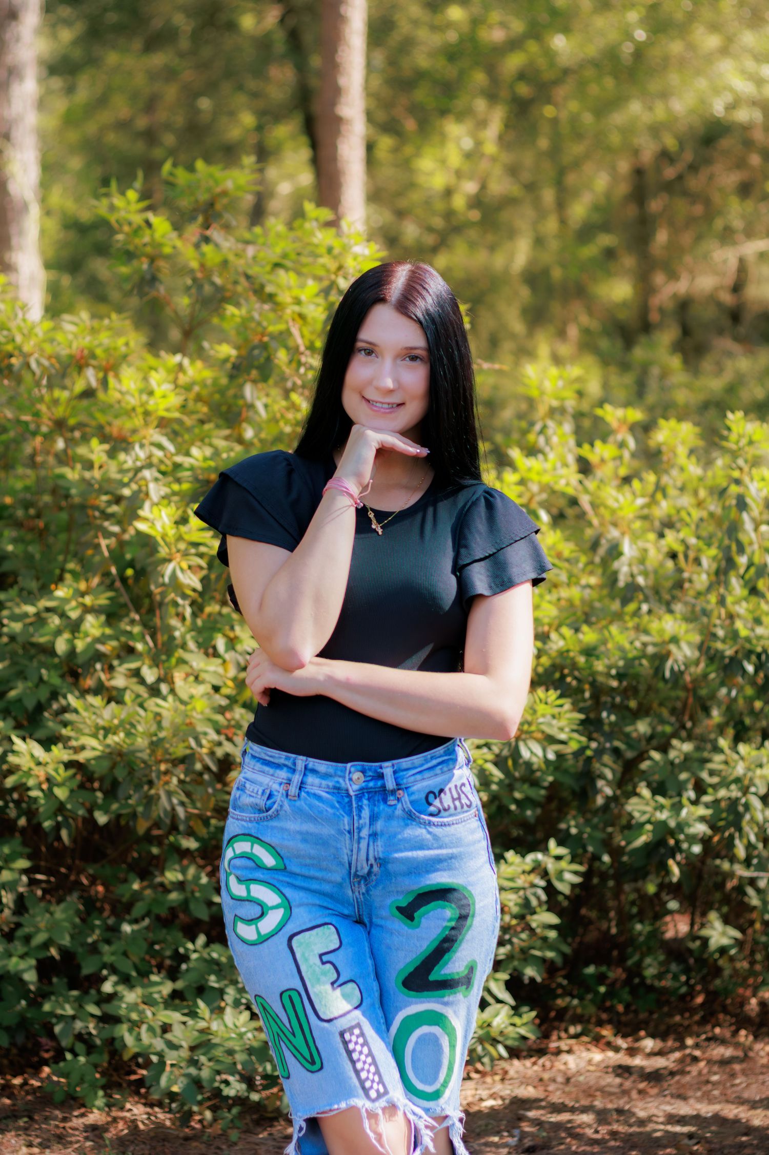 Young woman with black hair in black top and senior 2020 jeans poses outdoors among green trees.