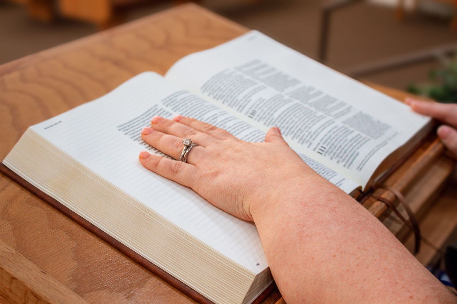 Close-up of woman's hand with ring resting on open Bible on wooden table.