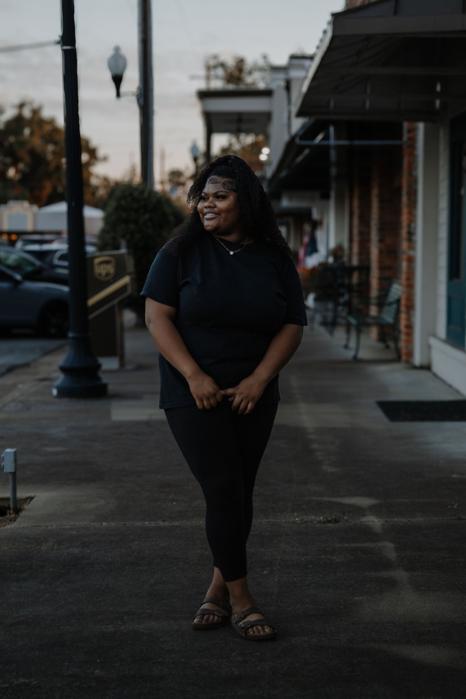 Young Black woman in all-black outfit smiles while walking along a small town sidewalk at dusk.