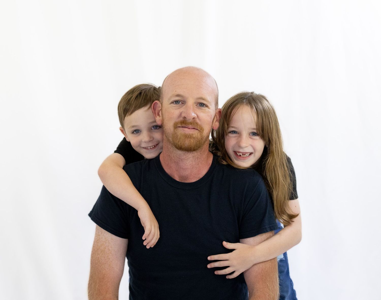 Bald man with red beard in black shirt hugging two young children smiling against white background.