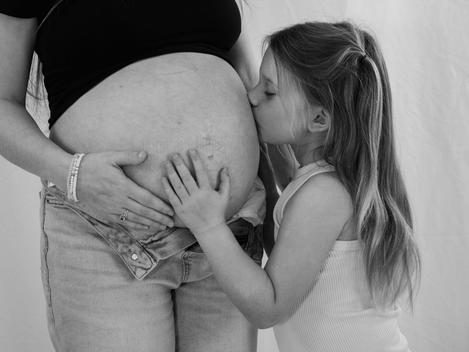 Black and white photo of young girl kissing a pregnant womans bare baby bump affectionately.
