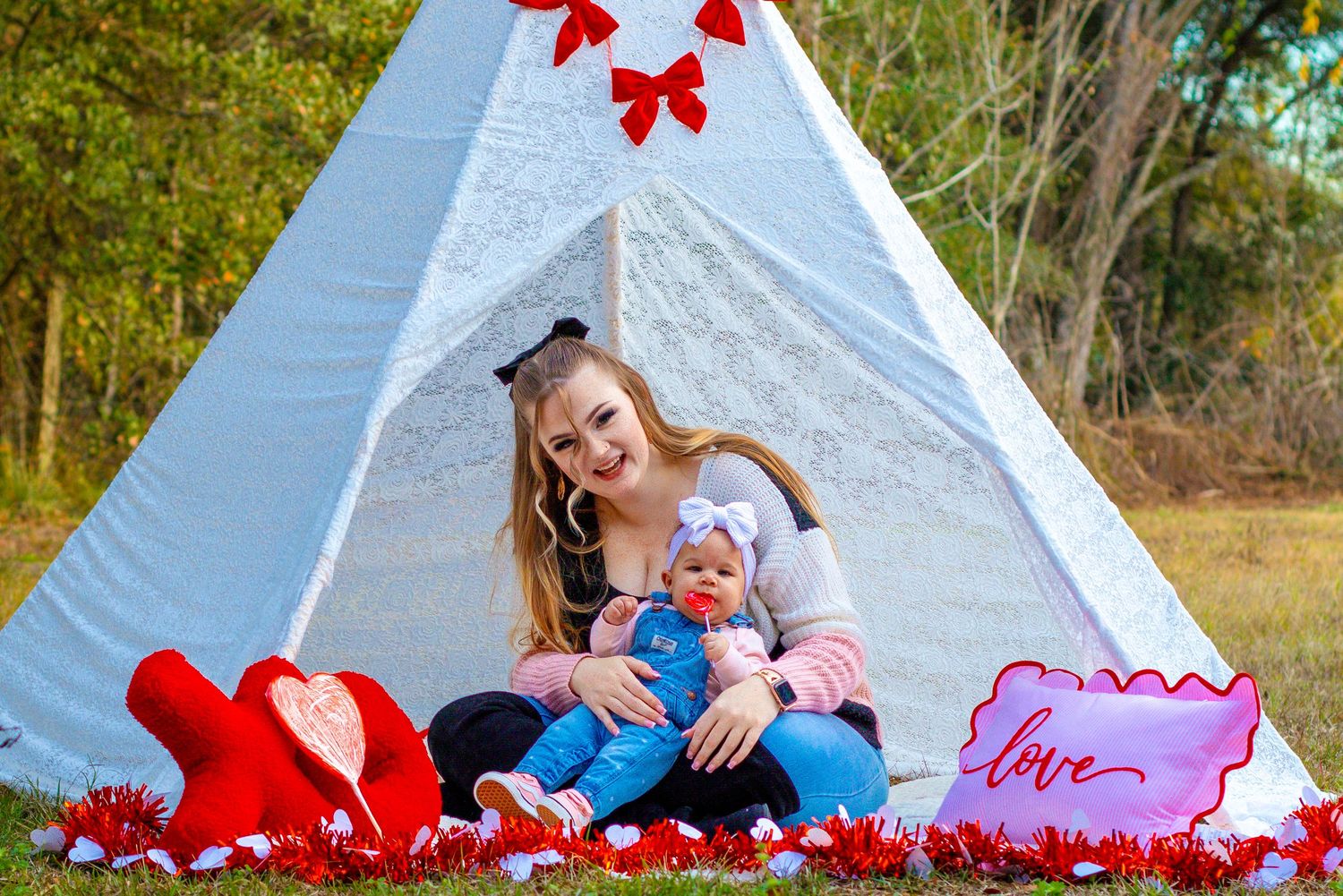 Smiling woman holds baby doll while sitting in front of white teepee surrounded by red Valentine decorations.