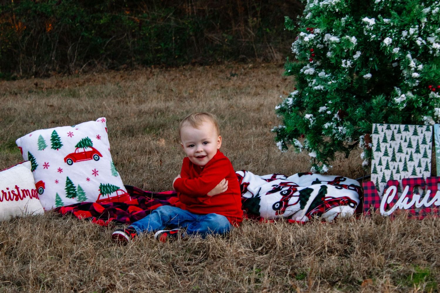 A smiling toddler in red sits on a plaid blanket beside a snowy Christmas tree outdoors.