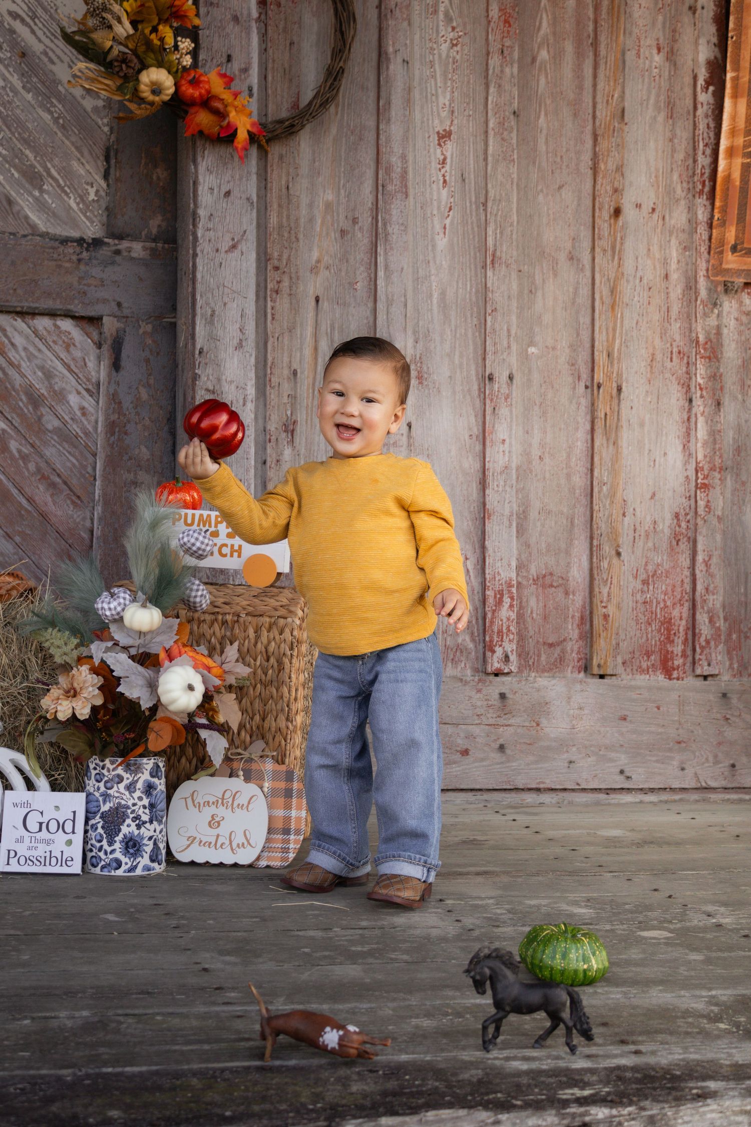 Laughing toddler in yellow sweater holding red pumpkin near fall harvest barn decorations.