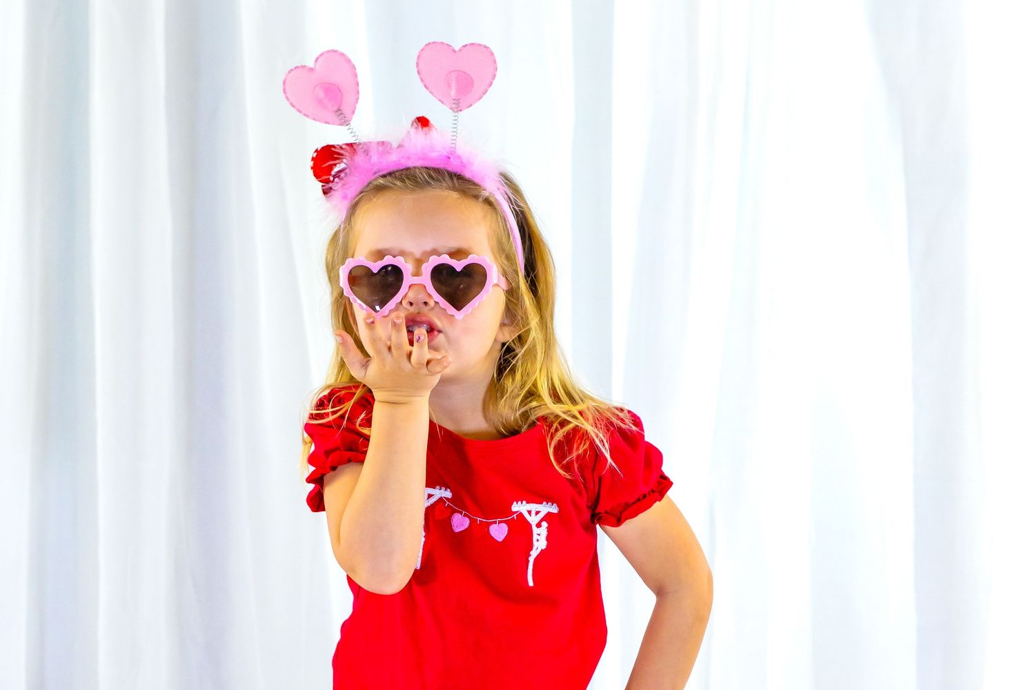 Little girl in red dress wears heart-shaped sunglasses and pink heart headband while blowing a kiss.