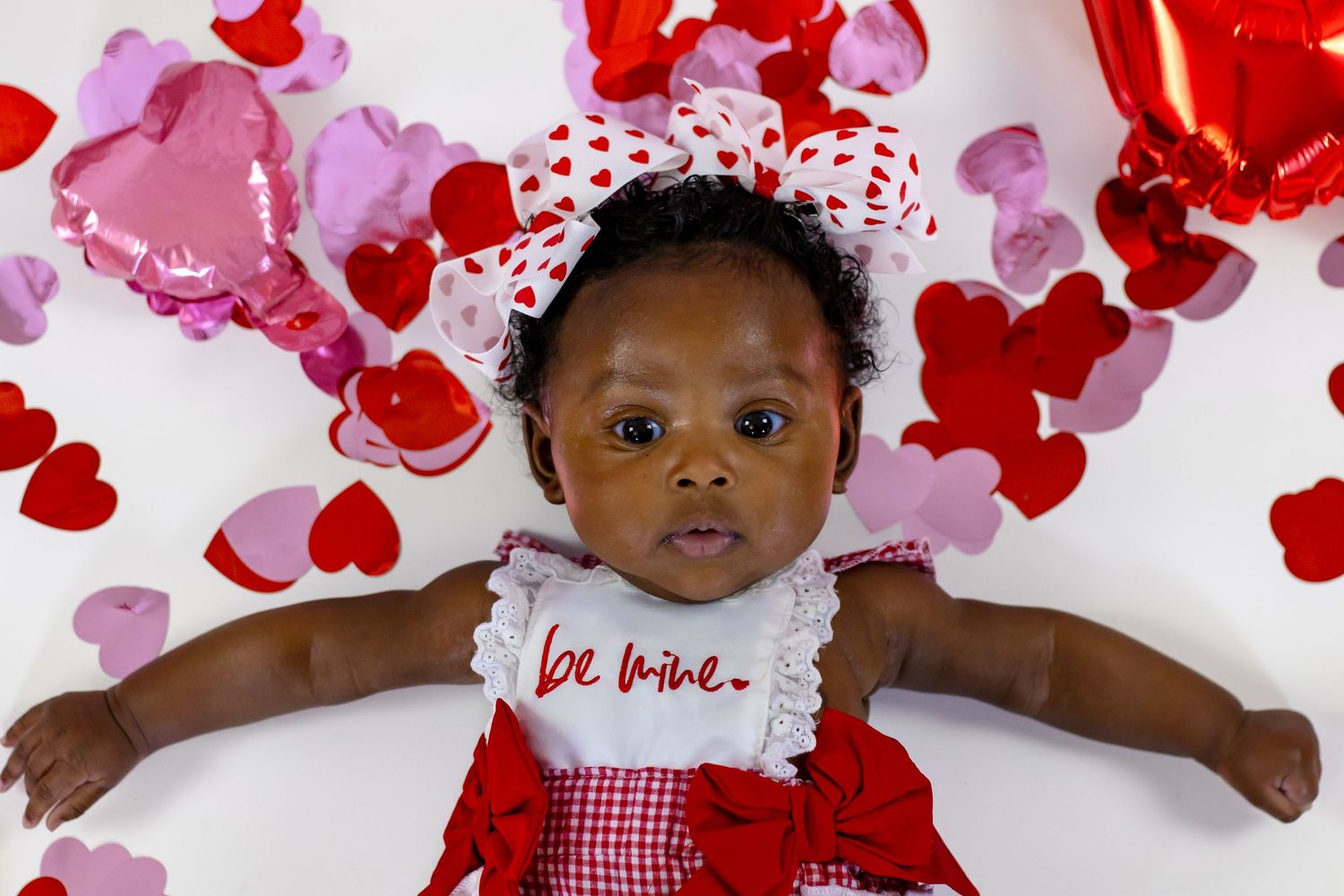 Close-up of baby in red gingham be mine dress surrounded by colorful heart confetti for Valentine's Day.