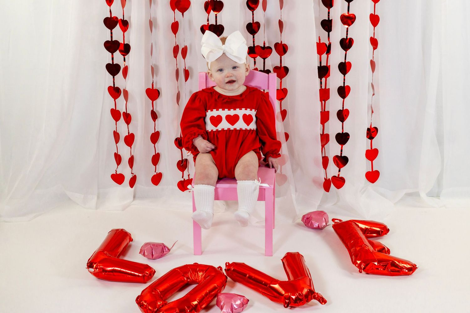 Baby girl in red heart romper sitting in Valentine's Day decorated studio with LOVE balloons.