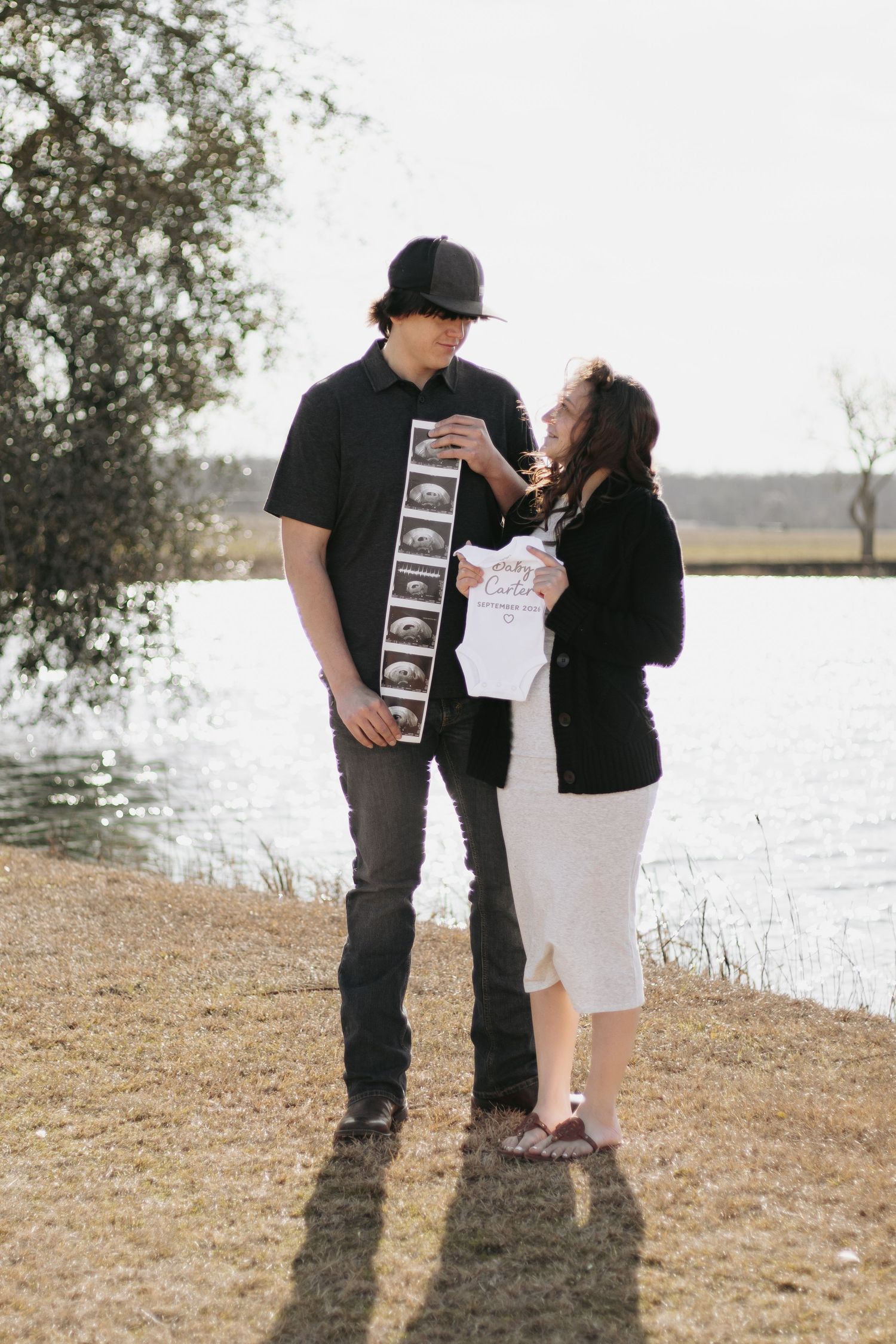 Young couple shares a tender moment holding ultrasound photos and baby onesie by a lake for pregnancy announcement.