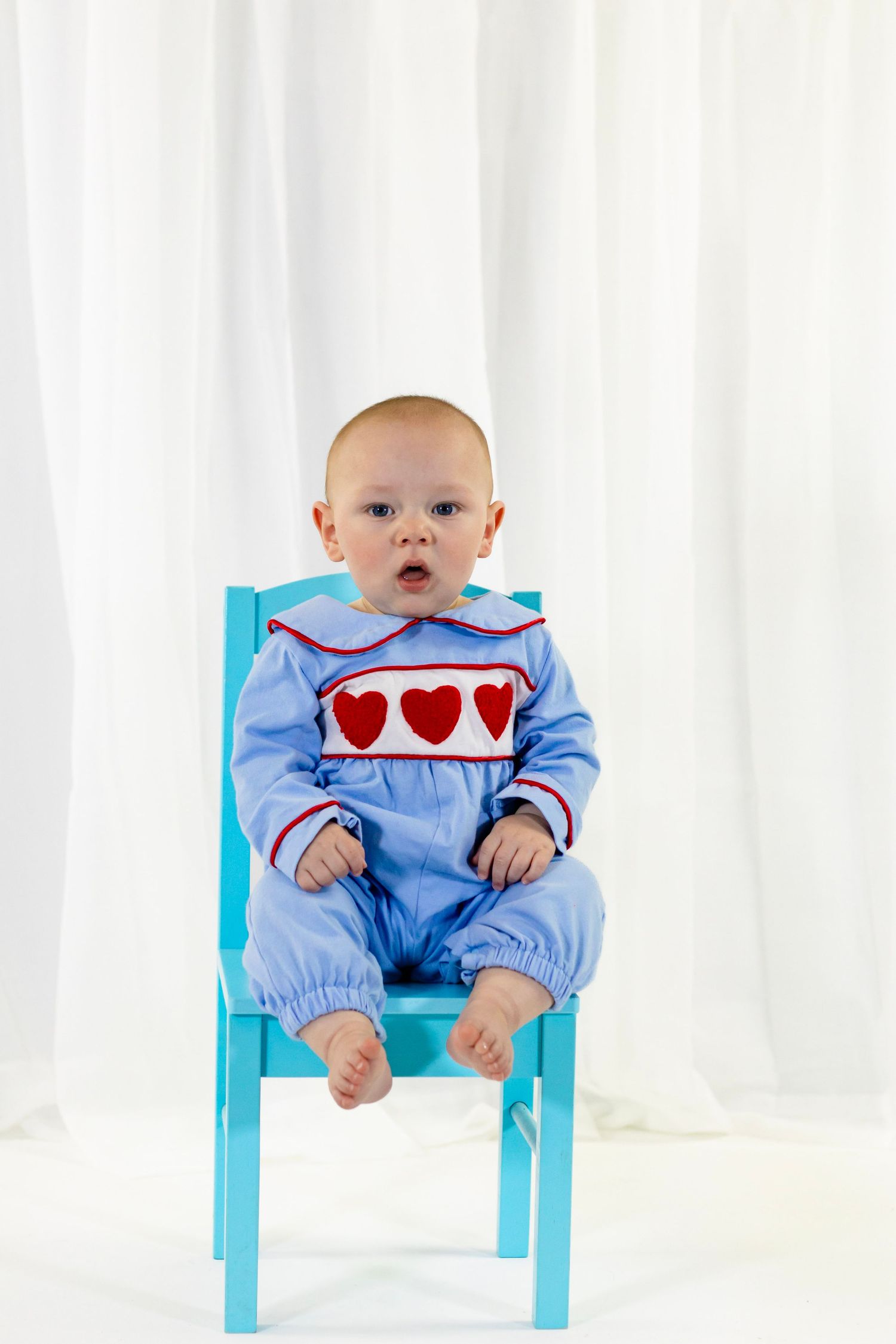 Baby in blue smocked heart romper sits on a teal chair against white curtain backdrop for Valentine's Day.
