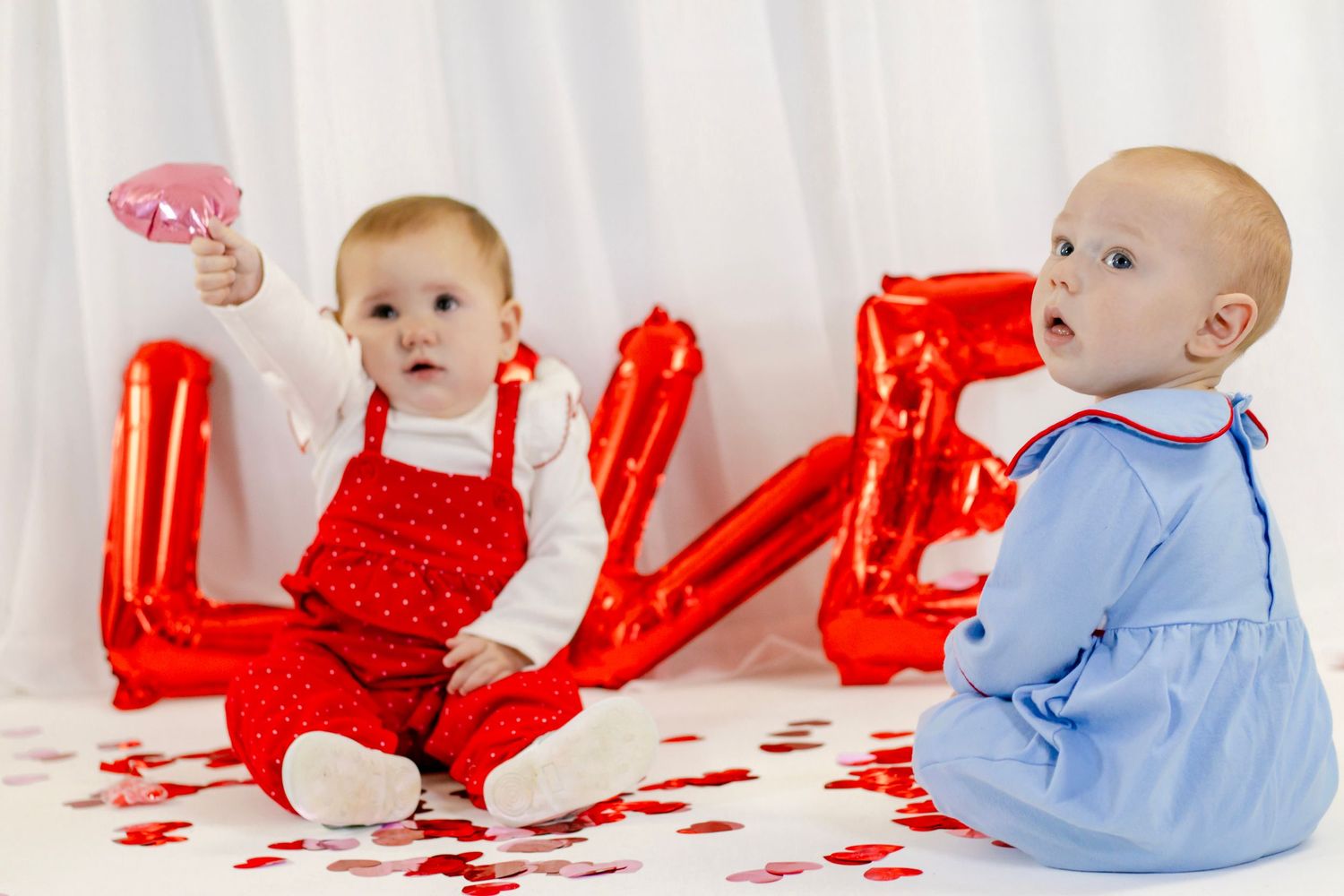 Two babies in red and blue Valentine outfits sit before red LOVE balloons on a white background.