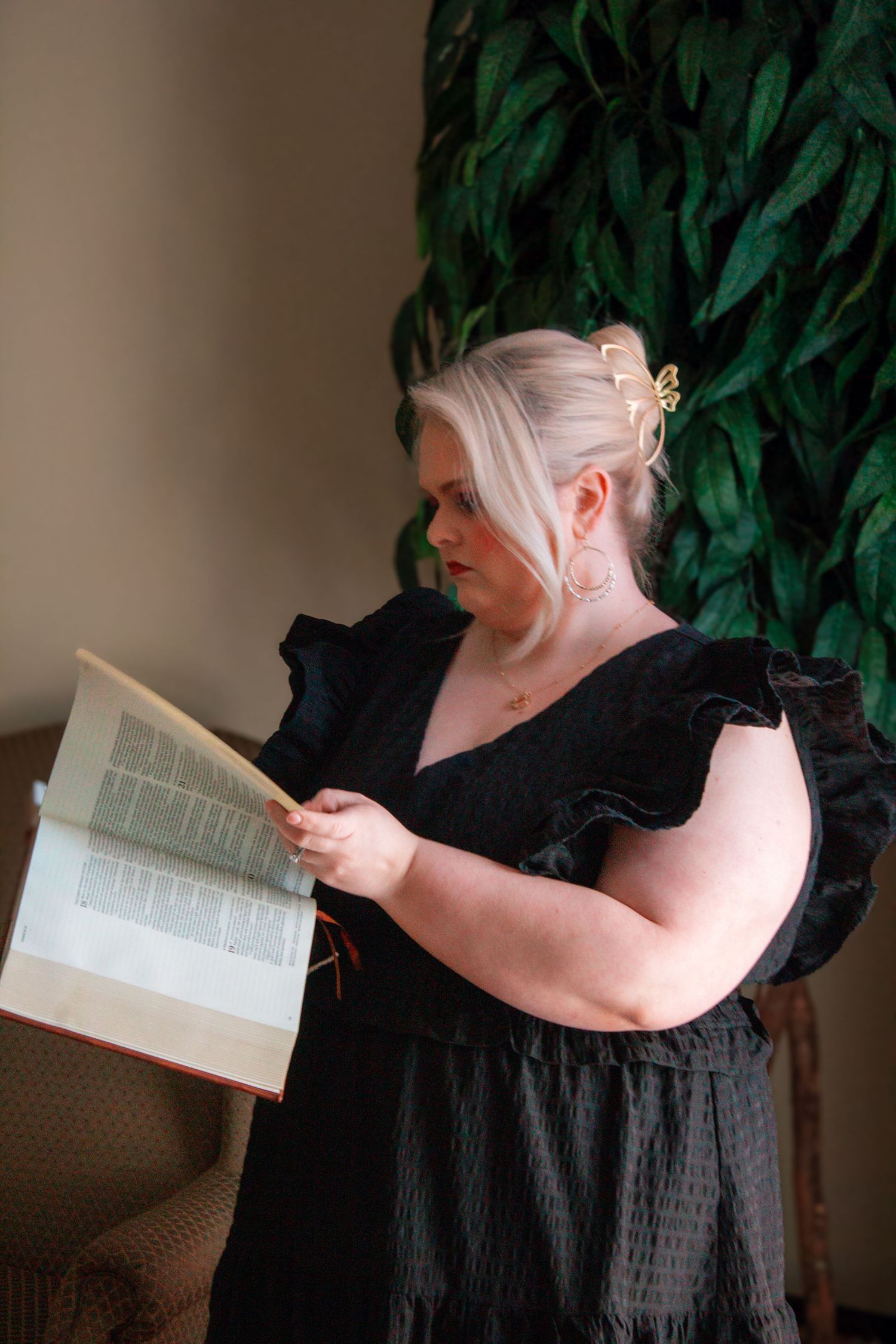 Blonde woman in black ruffled dress reading an open book while standing near a leafy green plant.