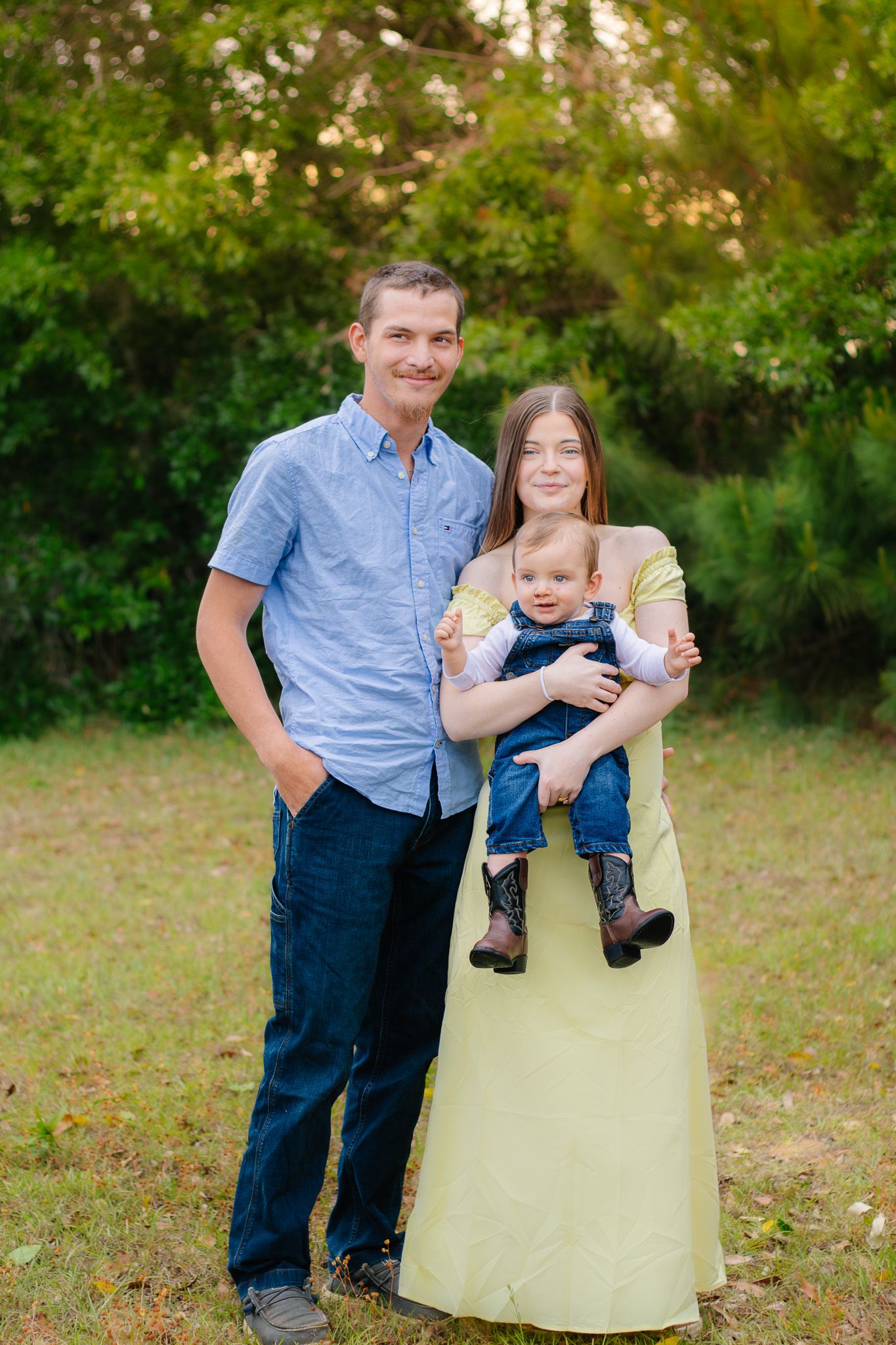 Young family of three posing outdoors, parents smiling with baby in denim overalls, lush green trees background.