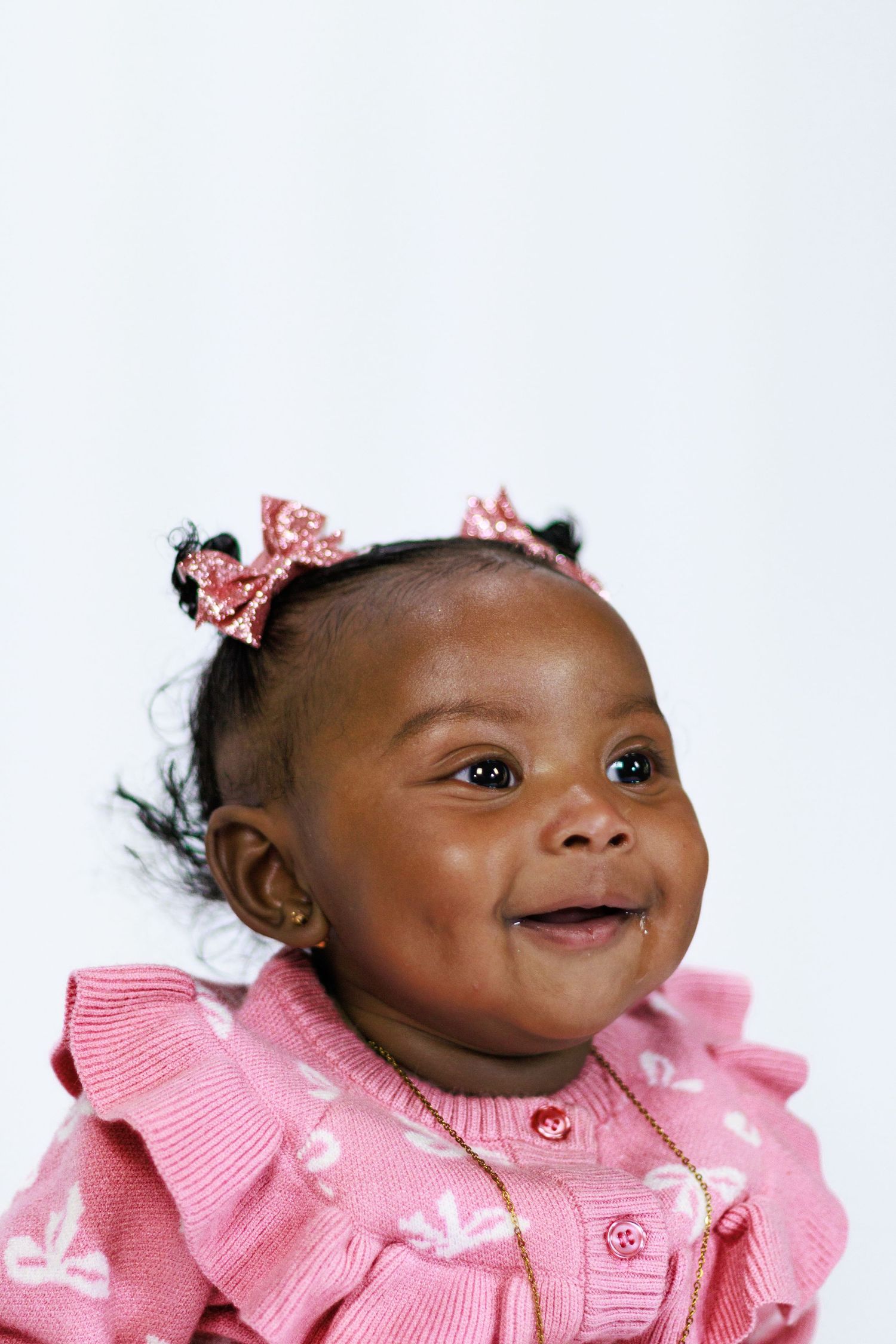 Smiling African American baby girl wearing a pink sweater and bow hair accessories against a white background.