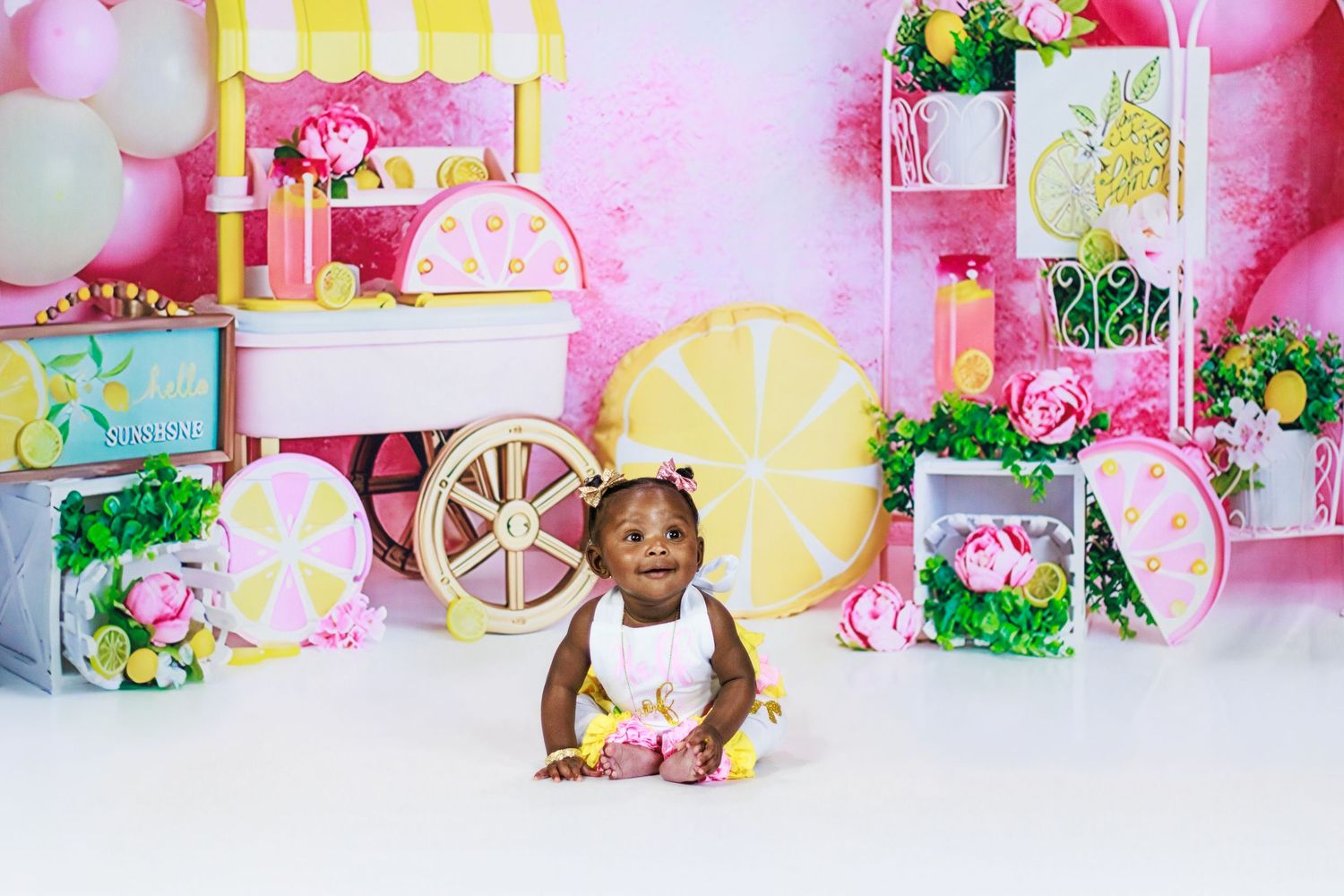 Adorable baby girl in a white lemon dress sitting in front of a vibrant pink lemonade stand birthday backdrop.
