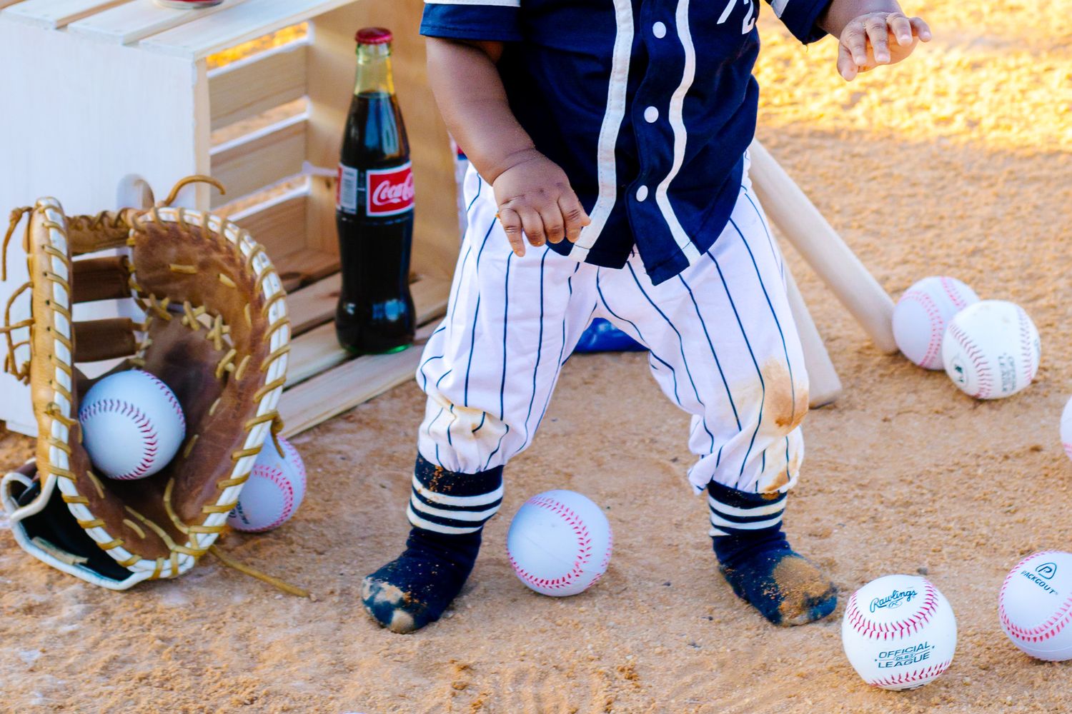Baby in pinstripe baseball uniform standing near glove, balls, and Coca-Cola bottle.