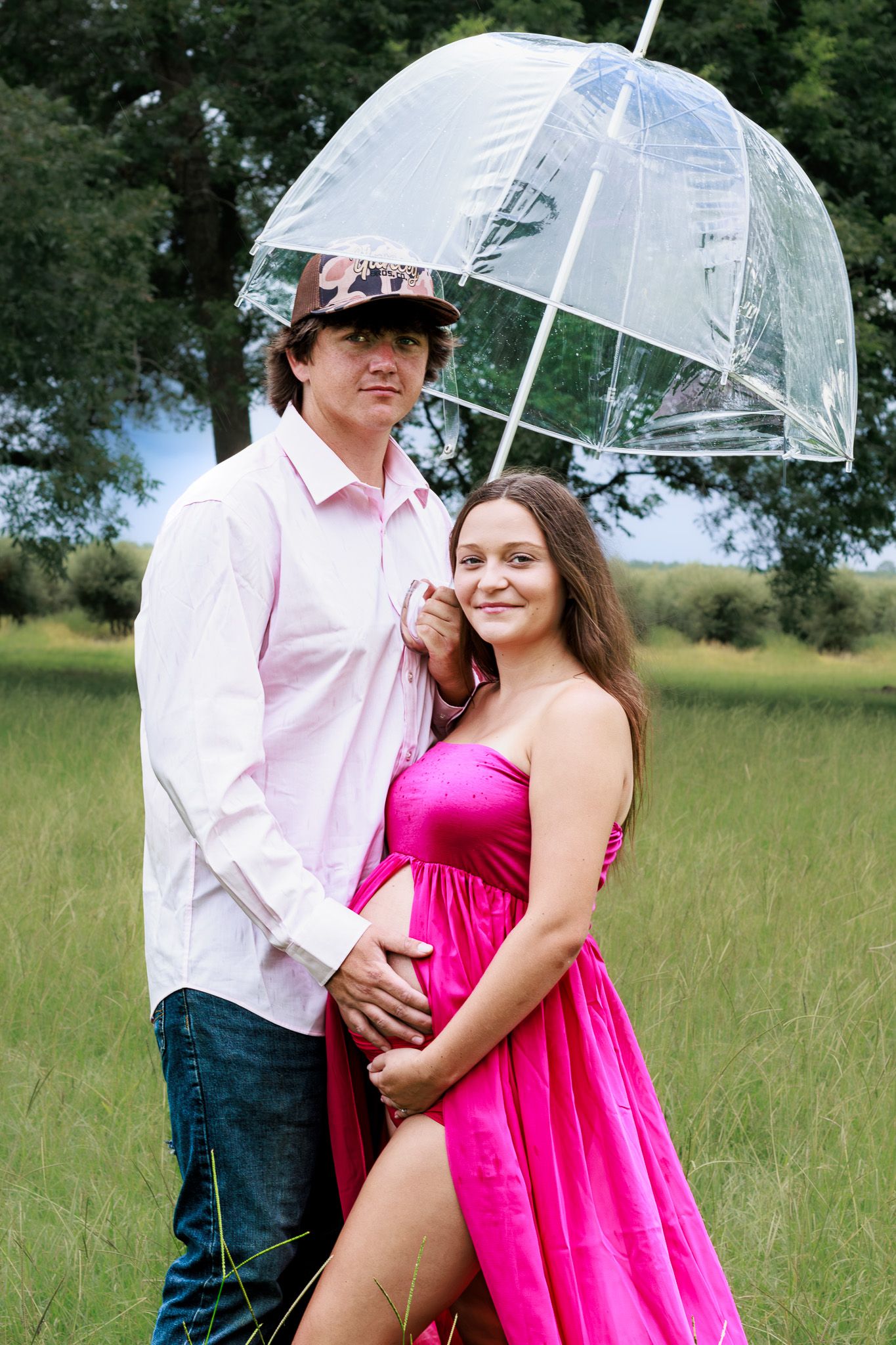 Expecting couple standing under a clear bubble umbrella in a green field, woman in pink maternity dress.