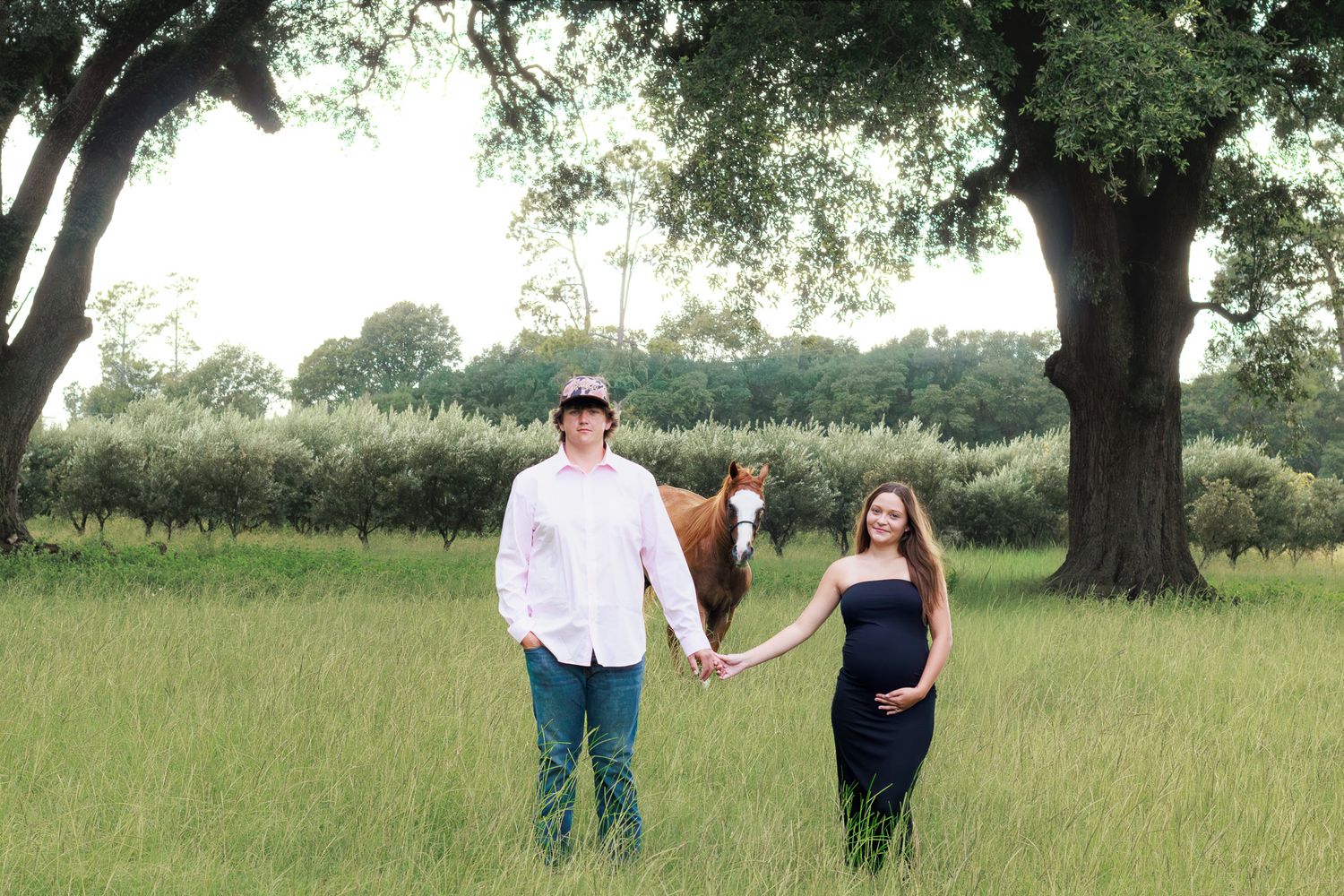 Couple holding hands in green field with horse and trees during outdoor maternity photo session.