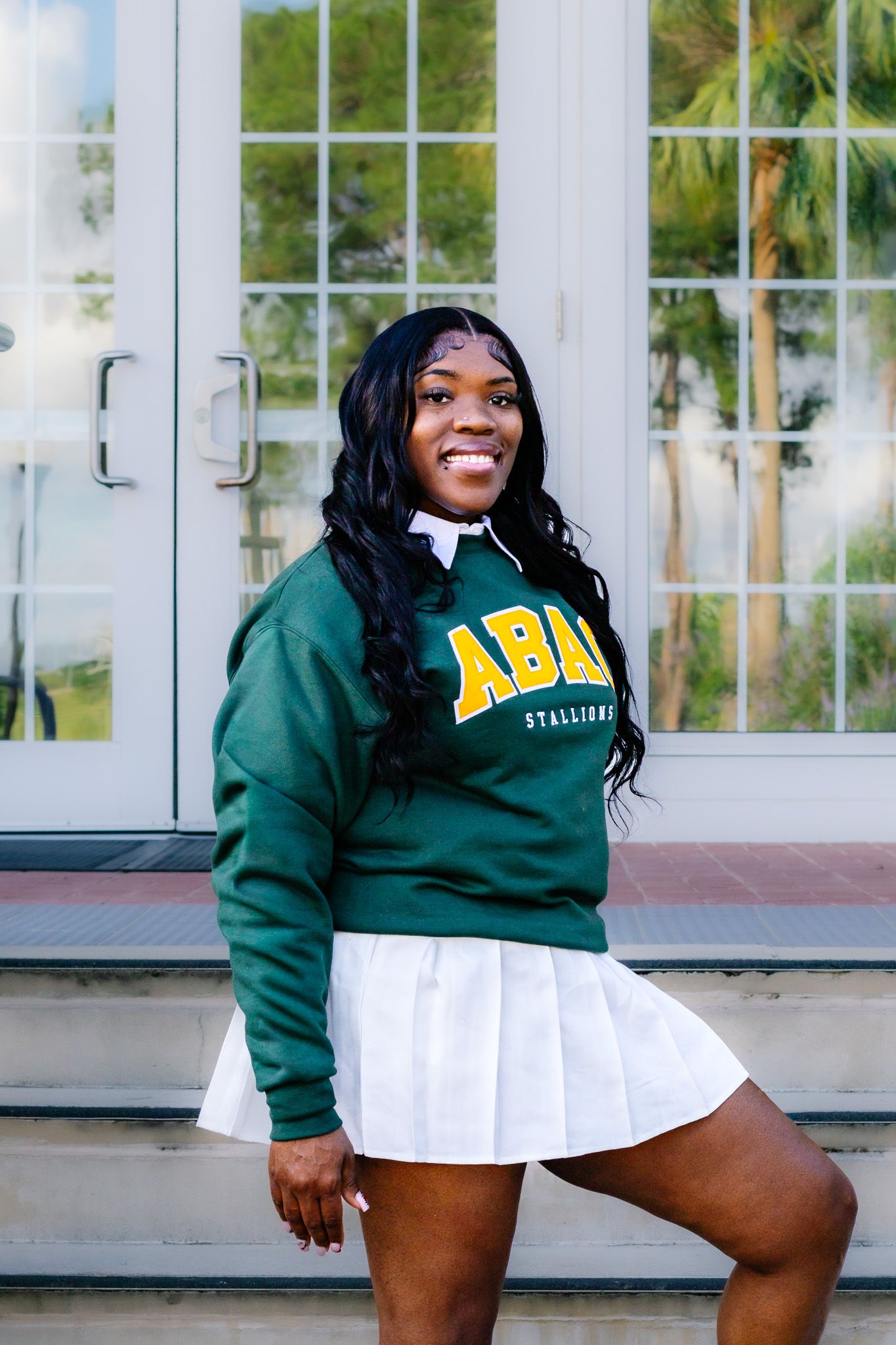 Smiling young woman wearing a green ABAC Stallions sweatshirt and white pleated skirt posing outdoors.