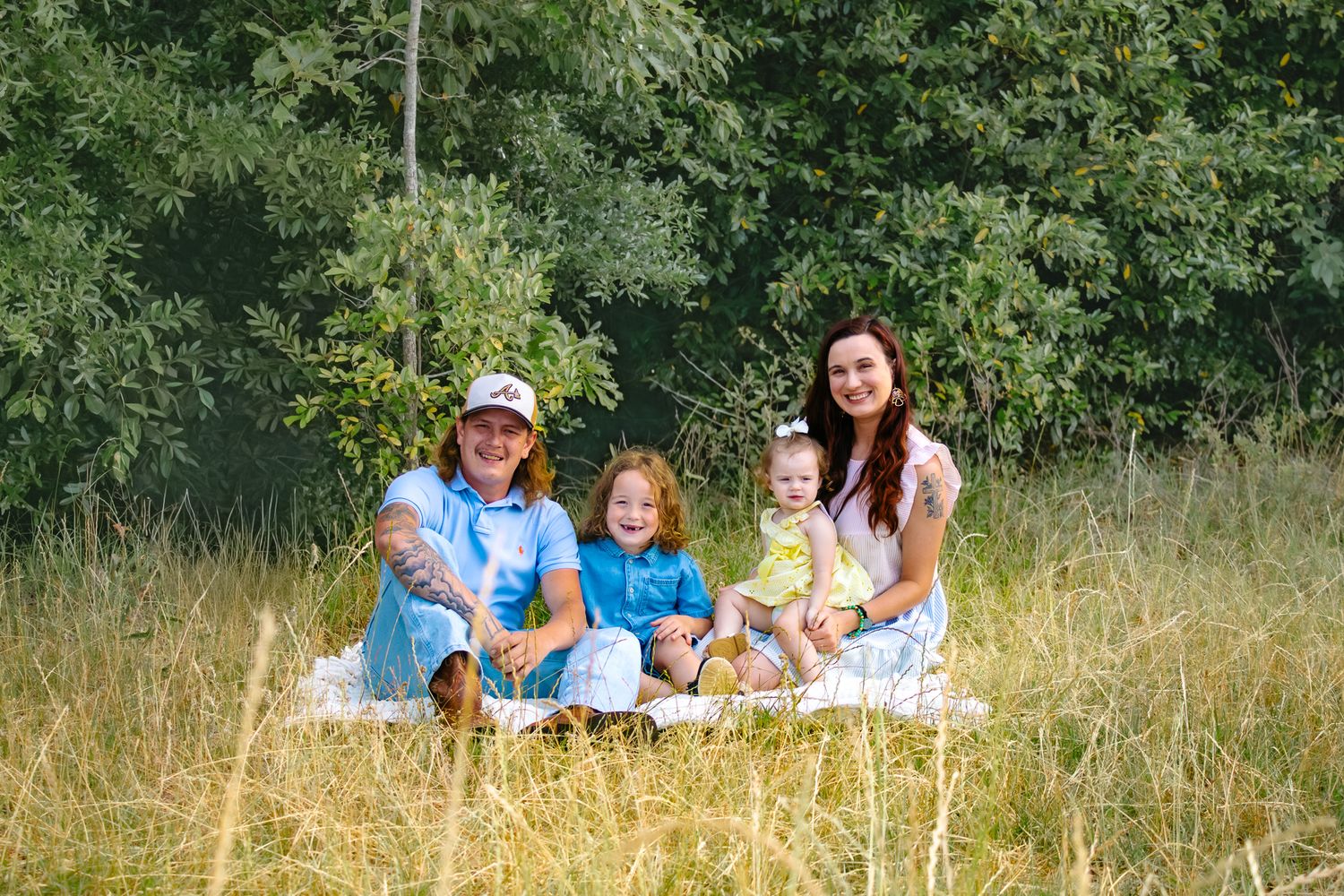 Family of four sitting in tall grass field with lush green trees, two adults and two young children in matching blue outfits.