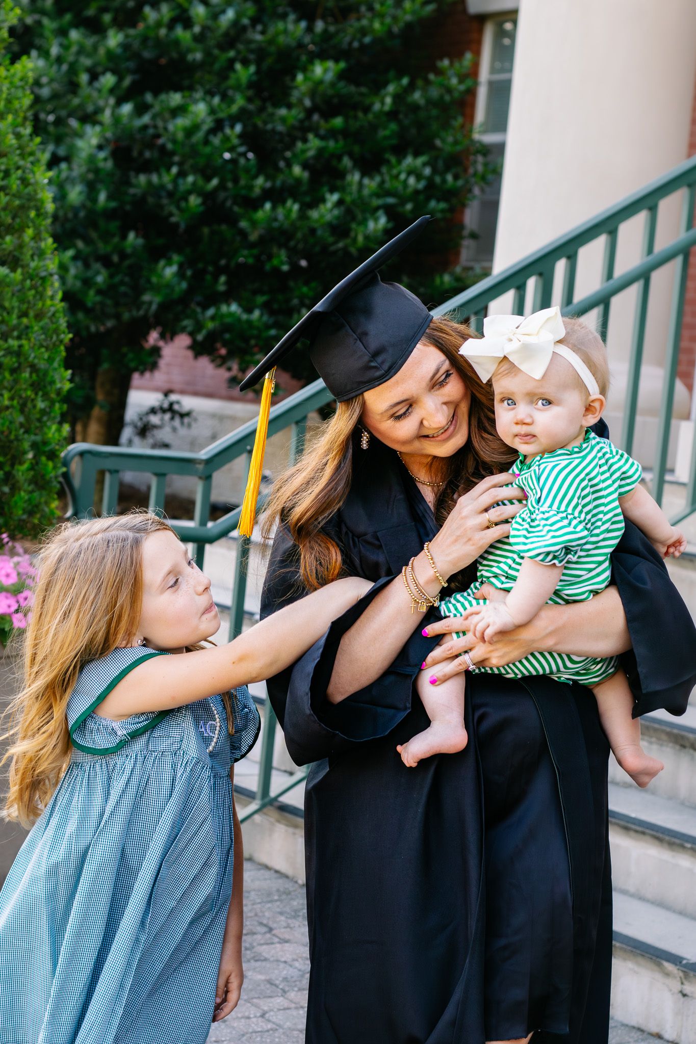 Graduate in cap and gown holds baby while young girl reaches up during outdoor graduation celebration.