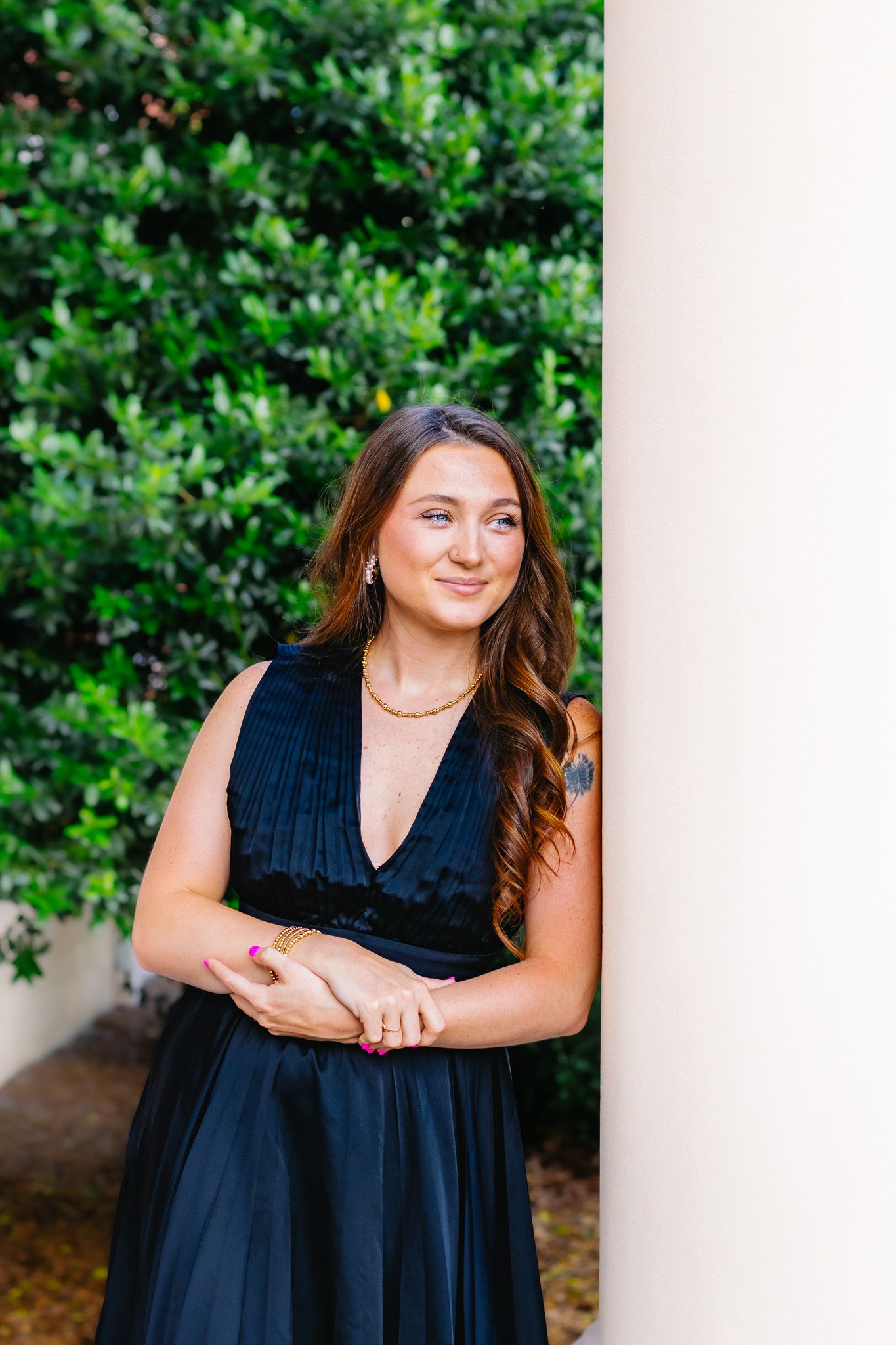 Young woman in elegant black dress leaning against white column with lush green foliage background.