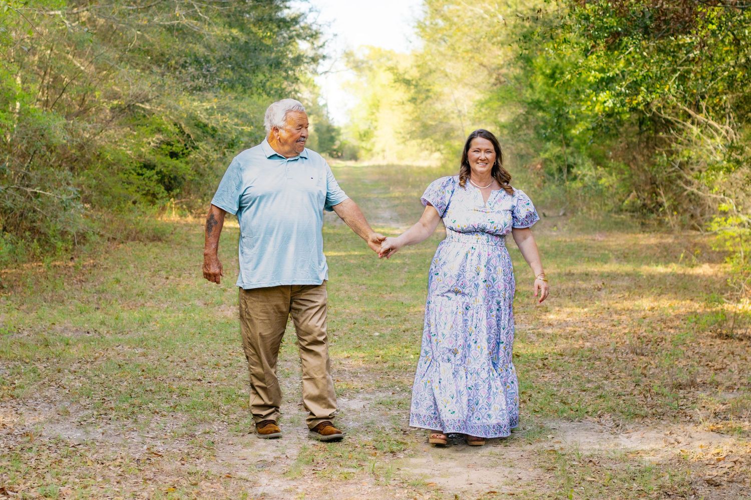 Couple holding hands walking on a sunlit dirt path surrounded by lush green trees.
