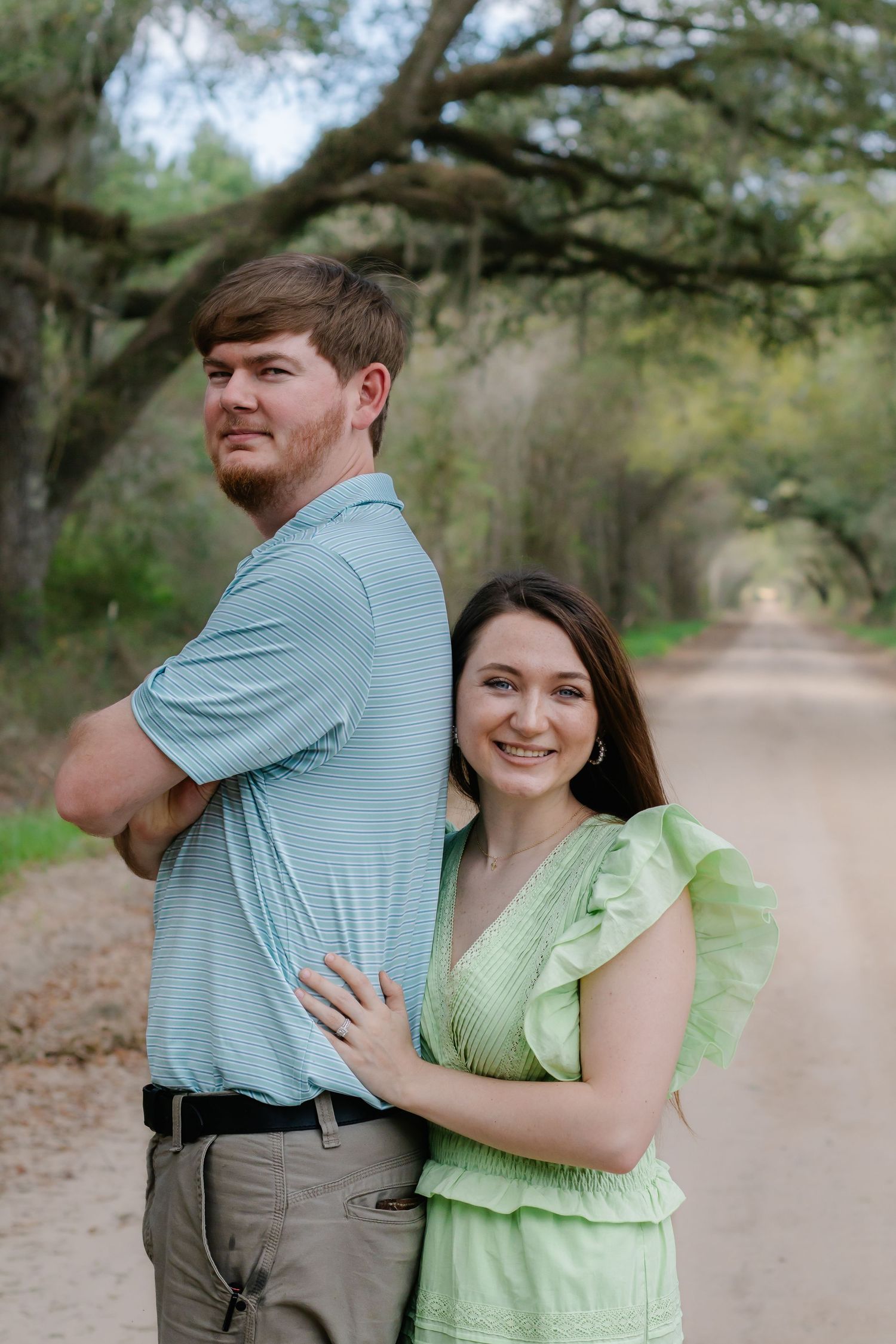 Smiling couple posing back-to-back on a scenic dirt road lined with moss-covered oak trees.