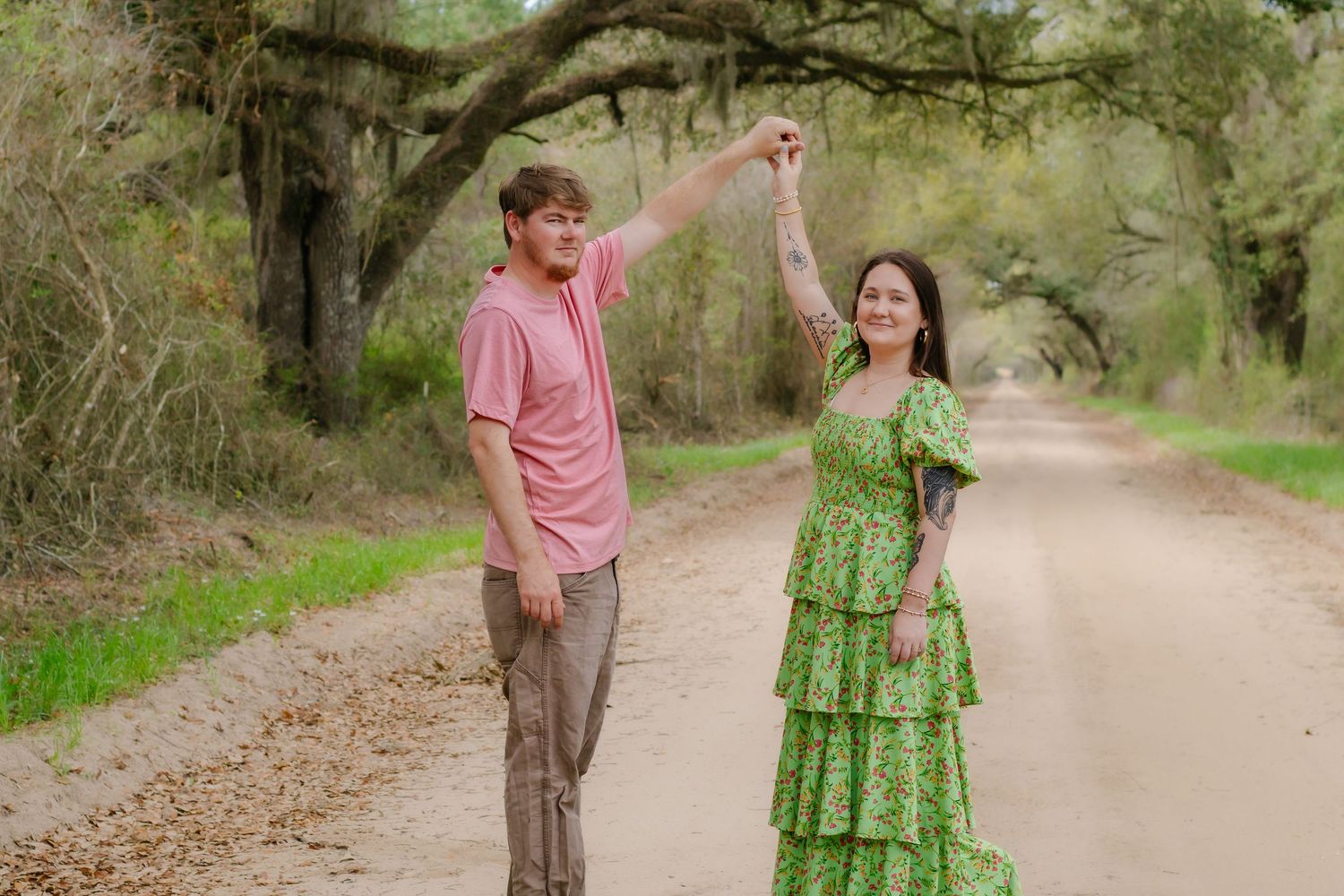Young couple dancing on a scenic tree-lined dirt road, man in pink shirt and woman in green dress.