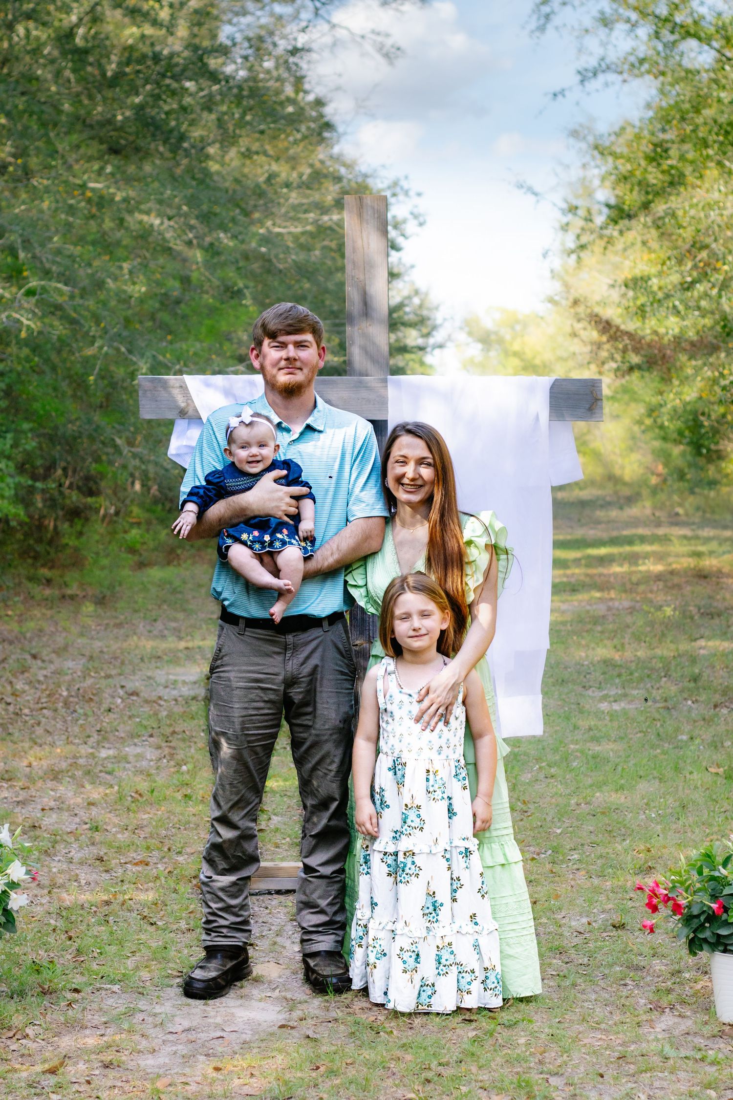 Young family of four standing before a large wooden cross with white draping in a lush outdoor setting.